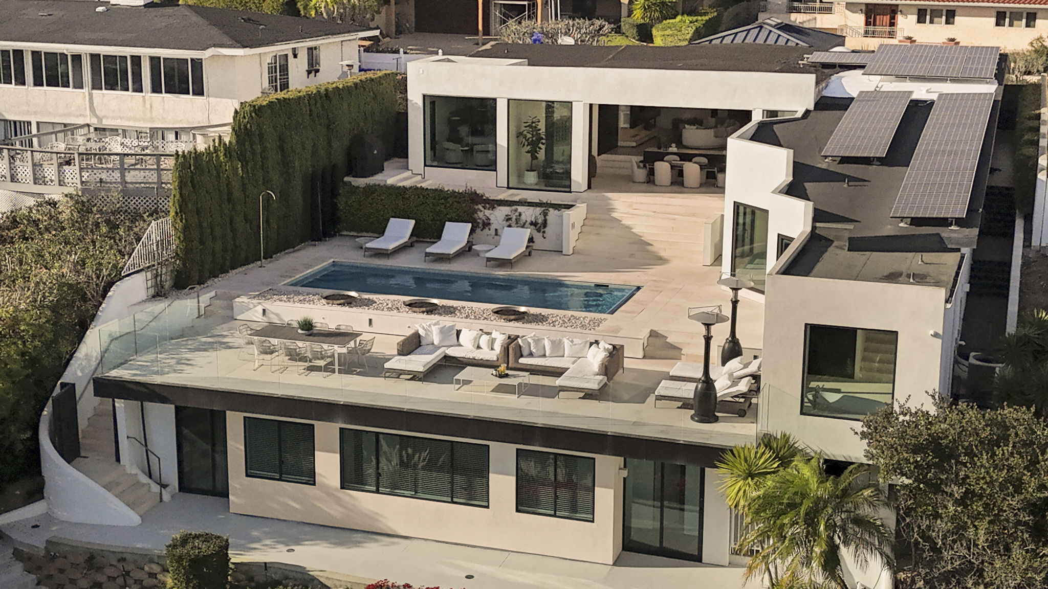 This is a high-perspective aerial view of a modern hillside residence with a prominent rear outdoor entertaining space. The property features a rectangular swimming pool, a spacious deck with contemporary lounge furniture, and integrated outdoor dining areas, all overlooking a multi-level terrace. The sleek architectural design incorporates large sliding glass doors, flat roofing with solar panels, and a refined ivory facade that creates a luxurious, resort-like atmosphere.