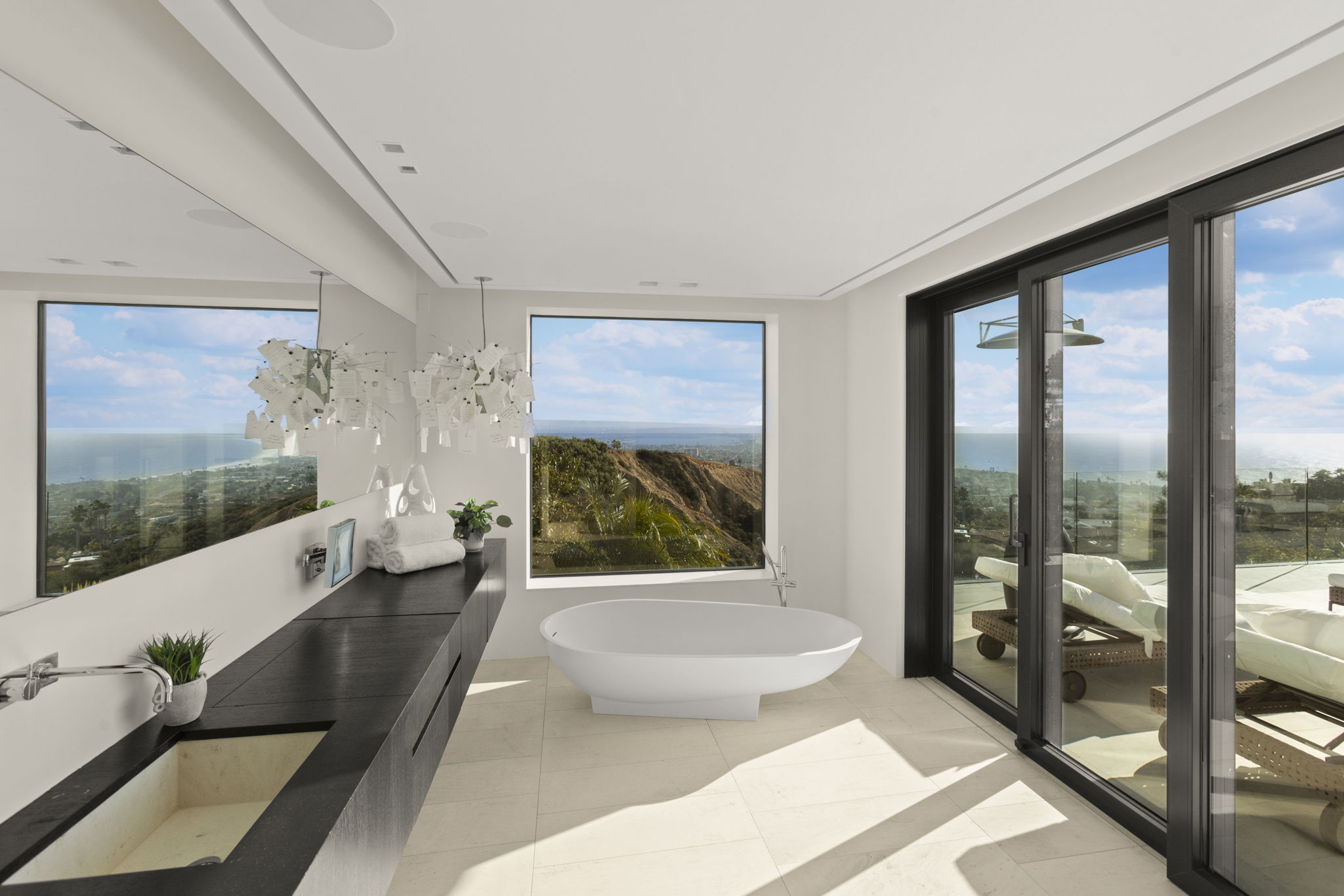 This luxurious primary bathroom features a sleek, minimalist aesthetic with a floating matte black vanity and a contemporary white soaking tub centered before a large picture window. The room is flooded with natural light and offers stunning, panoramic coastal views through both the floor-to-ceiling windows and the glass sliding doors that open to a private balcony. The clean lines, premium finishes, and integrated indoor-outdoor flow create a modern, high-end spa-like atmosphere.