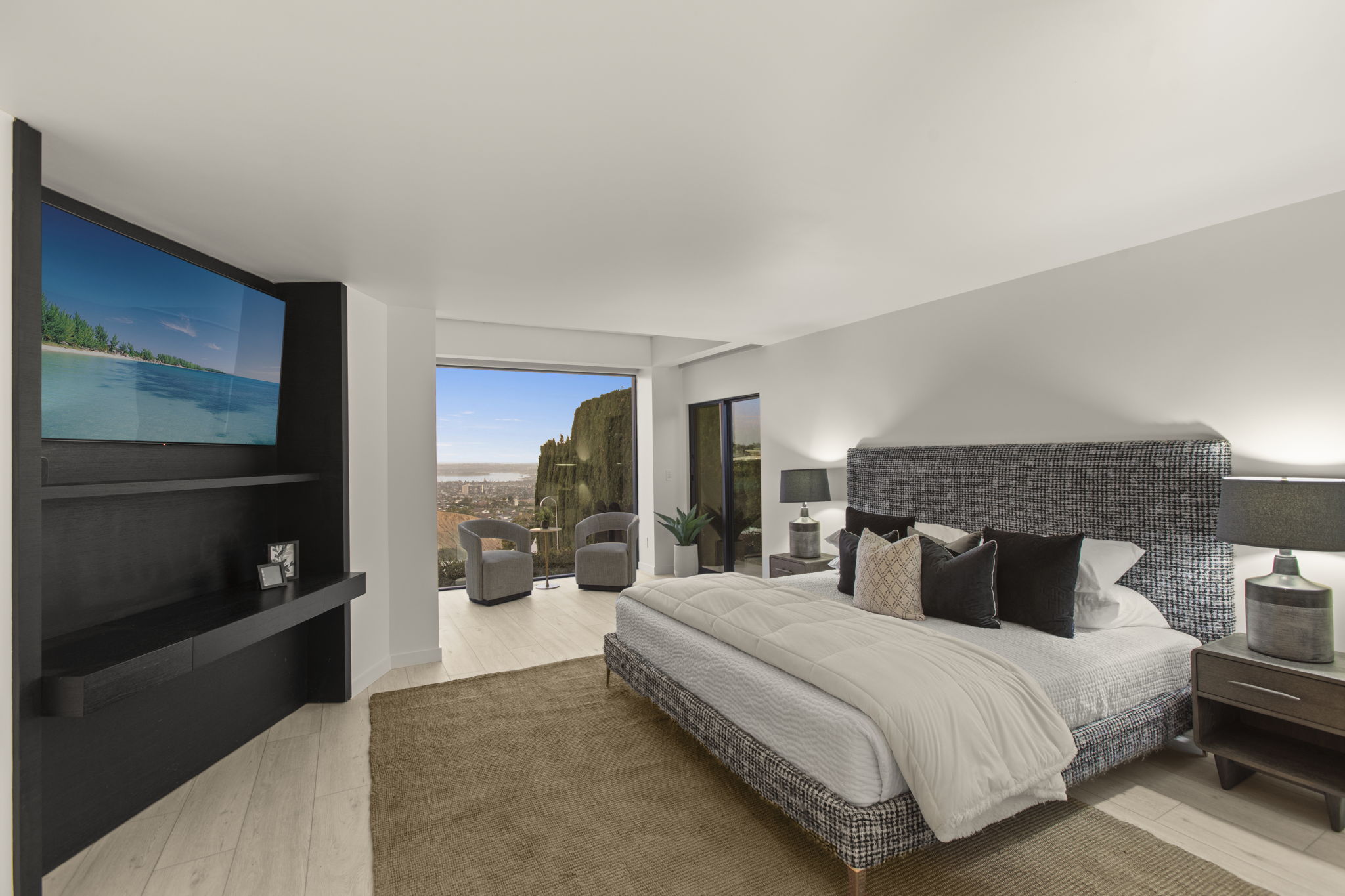 This luxurious primary bedroom features a modern, high-contrast design with a large, textured upholstered headboard and matching frame. The room is anchored by a spacious area rug and boasts a stunning floor-to-ceiling view of the hillside and city skyline from the attached private sitting area. Sleek built-in shelving with a wall-mounted flat-screen television adds convenience and contemporary flair to this elegant living space.