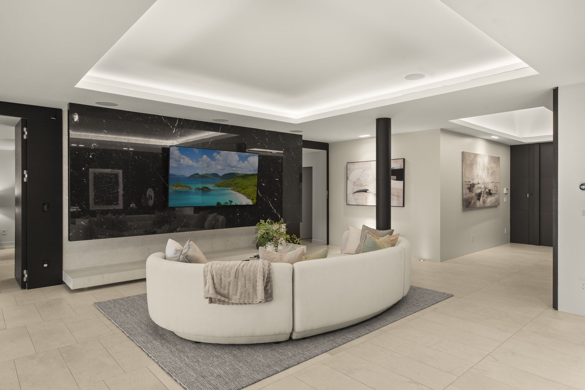 This modern living room features a striking black marble feature wall with an integrated television, offering a sophisticated focal point. A large, curved white sofa sits atop a neutral rug, creating a cozy and inviting seating area within the open-concept space. The room benefits from recessed cove lighting and minimalist decor, emphasizing a sleek, high-end contemporary aesthetic throughout.