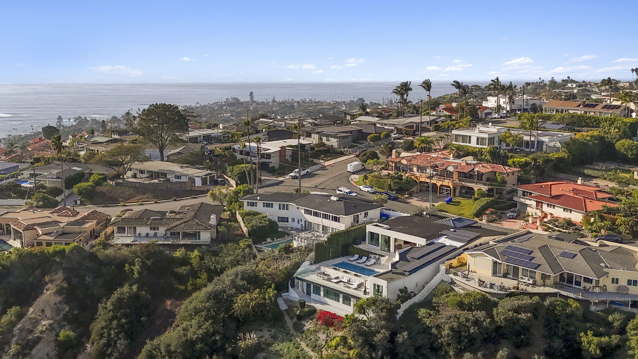 This high-angle, aerial perspective captures a picturesque coastal residential neighborhood with a variety of architectural styles, including modern and traditional homes. The image highlights the prominent elevated terrain featuring lush landscaping, private pools, and spacious decks, all set against a stunning backdrop of the ocean and horizon. It provides an excellent overview of the property's prime location, neighborhood layout, and expansive coastal exposure.