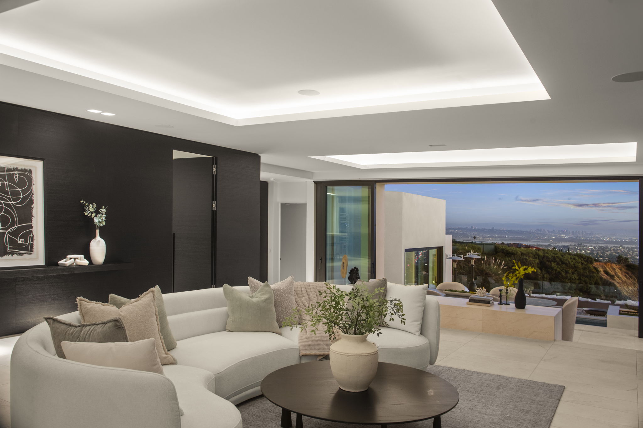 This sophisticated, contemporary living room features a expansive curved cream-colored sofa accented with plush pillows, centering around a round wooden coffee table. The sleek design is anchored by a dramatic dark-paneled feature wall and minimalist recessed ceiling lighting. Large glass slider doors seamlessly blend the interior with the terrace, offering stunning, unobstructed views of the sunset over the city landscape.