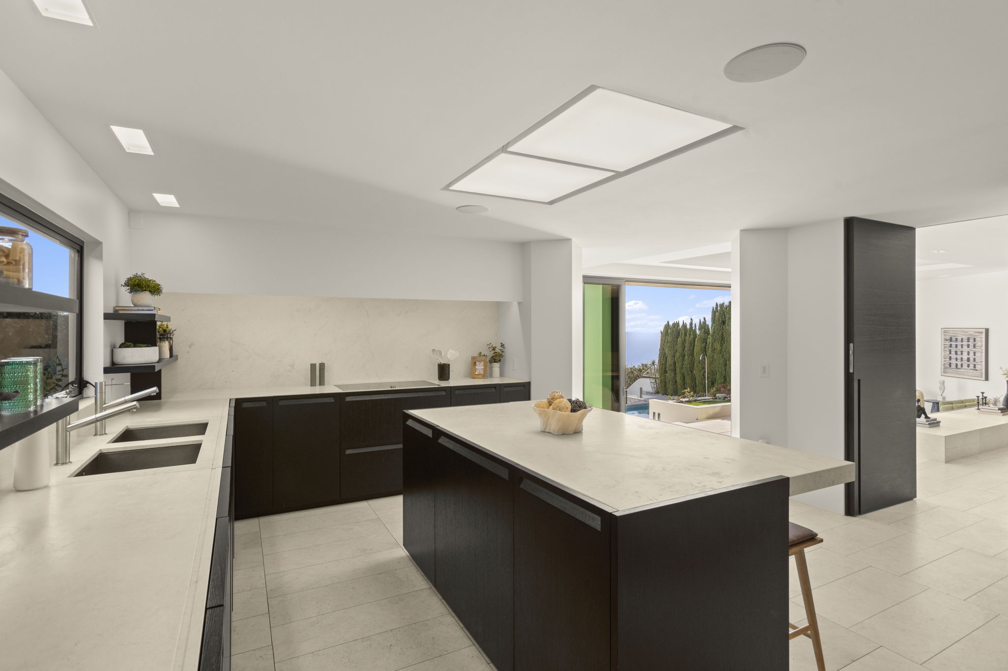 This sleek, contemporary kitchen features high-contrast dark wood cabinetry paired with light quartz countertops and a matching expansive center island. The open-concept design flows seamlessly into an outdoor patio area with scenic views, while clean lines and minimalist fixtures enhance the sophisticated, modern aesthetic.
