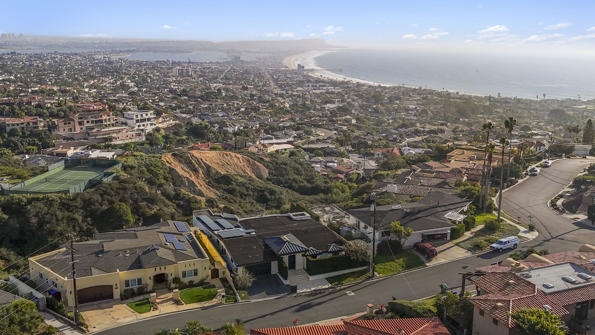 This stunning high-angle aerial view captures a coastal residential neighborhood nestled against a hillside, overlooking an expansive ocean coastline. The foreground features well-maintained, single-story homes with tile roofs and driveways, while the surroundings showcase lush vegetation, a local tennis court, and sweeping panoramic views of the distant shore. The imagery conveys a premium, active lifestyle location characterized by high elevation and proximity to the Pacific coast.
