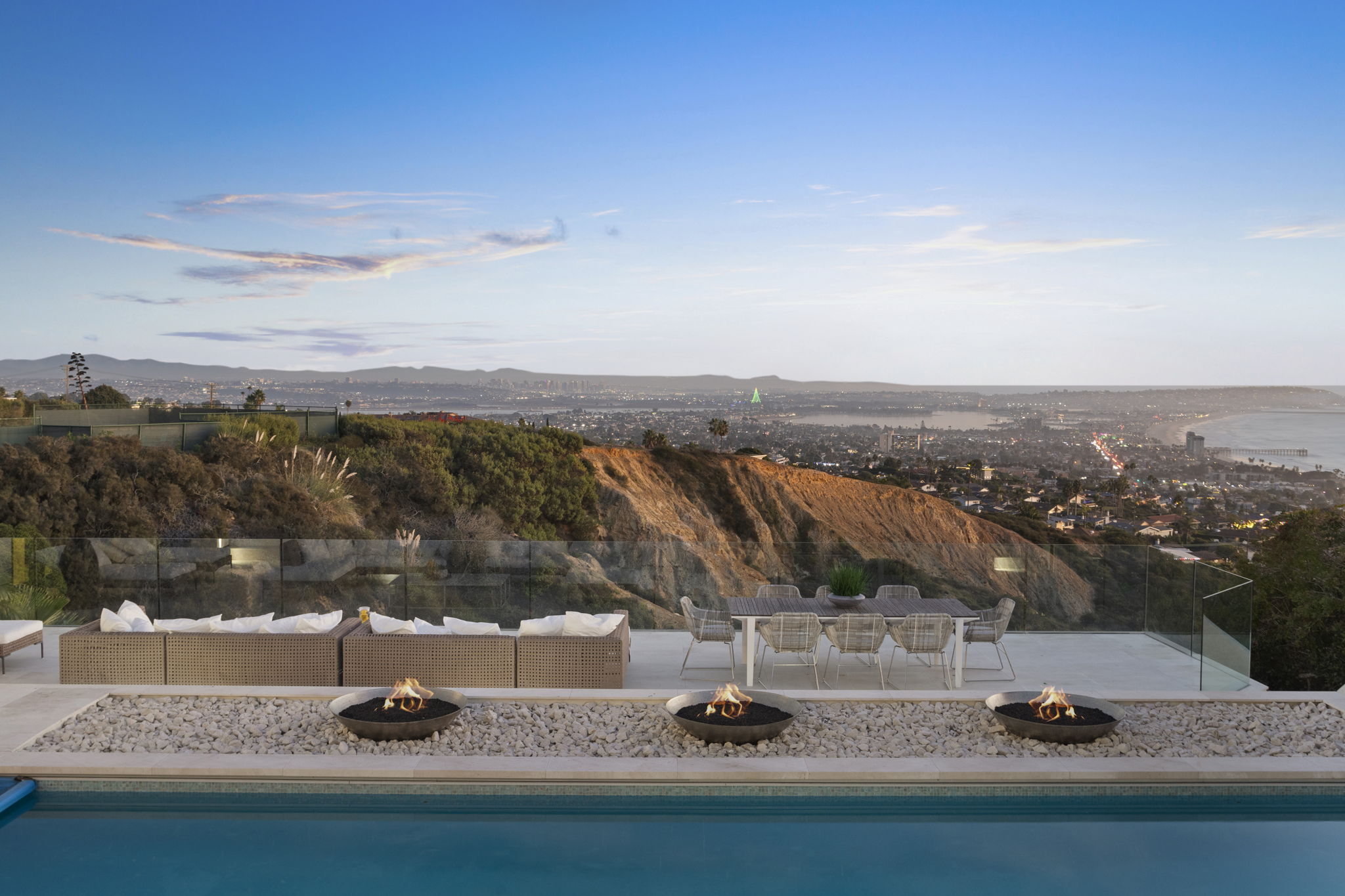This stunning outdoor space features a luxurious patio area equipped with comfortable lounge seating and a dining table, perfect for entertaining against a breathtaking panoramic coastal background. A sleek glass railing offers unobstructed views of the cliffs, city lights, and ocean, while three contemporary fire bowls line the edge of a pristine infinity pool, creating a dramatic and high-end atmosphere.