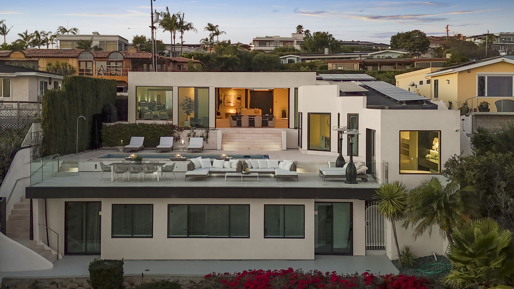 This modern, multi-level coastal home features an expansive deck with a swimming pool, outdoor seating areas, and dual fire pits, perfect for indoor-outdoor entertaining. Large sliding glass doors open the main living space to the terrace, emphasizing a seamless flow and impressive views. The architecture is clean and contemporary, accentuated by soft evening lighting and mature landscaping.