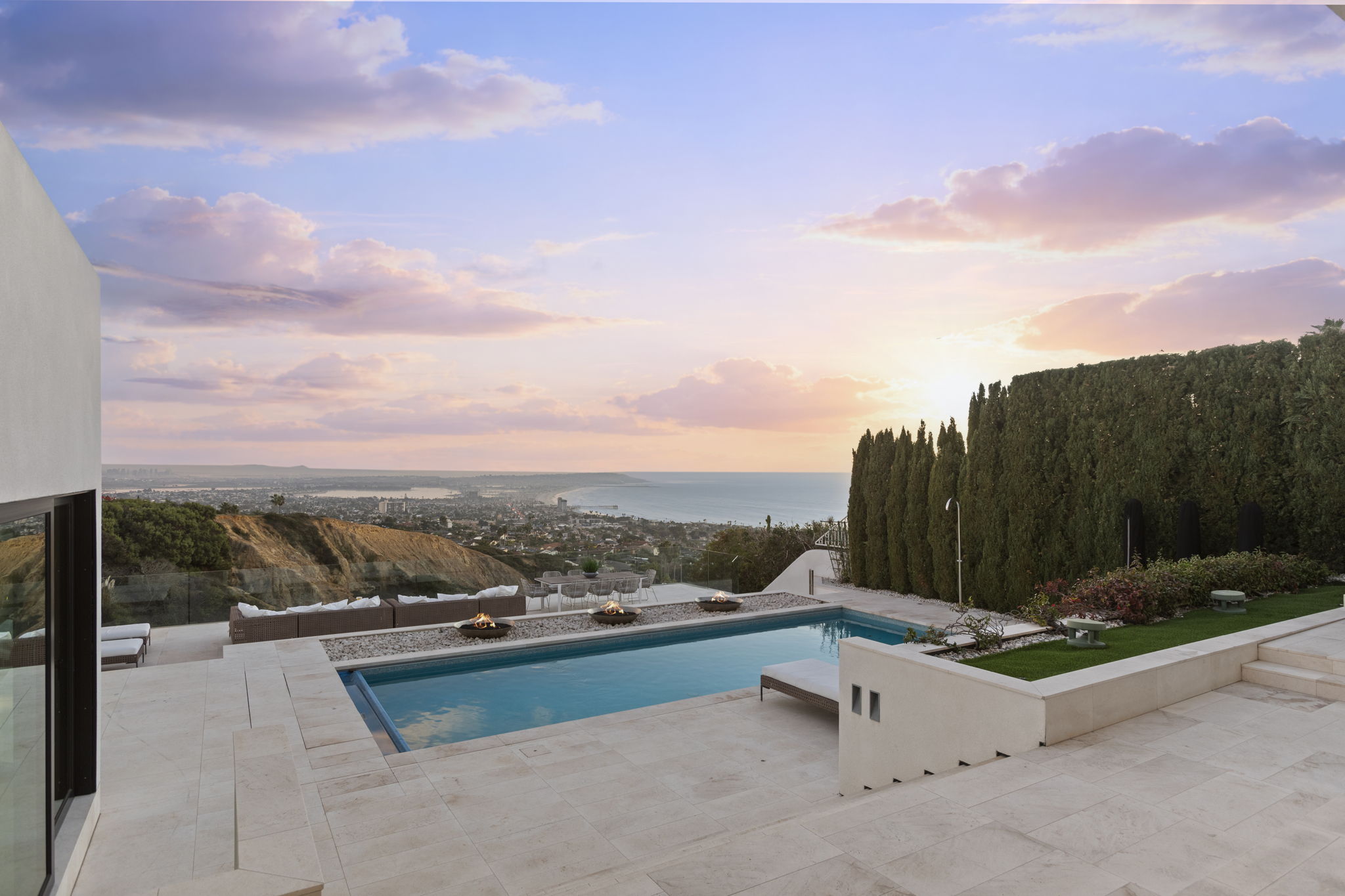 This stunning terrace features a rectangular infinity-edge pool overlooking a panoramic coastal view at sunset. The outdoor space includes curated landscaping, elegant fire bowls, a modern patio area with outdoor seating, and a sleek, contemporary design aesthetic. Ideally suited for luxury entertaining, the area perfectly blends sophisticated architecture with the natural beauty of the surrounding environment.