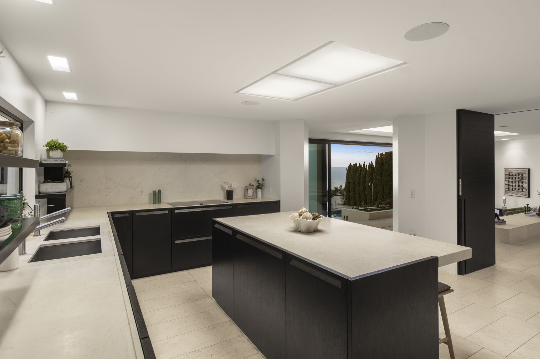 This sleek, contemporary kitchen features high-contrast dark wood cabinets paired with light stone countertops and matching backsplash. A large central island provides functional prep space, while an open floor plan seamlessly connects the kitchen to the living area and an outdoor view. Recessed lighting accents the clean, modern aesthetic, creating a sophisticated and airy environment.
