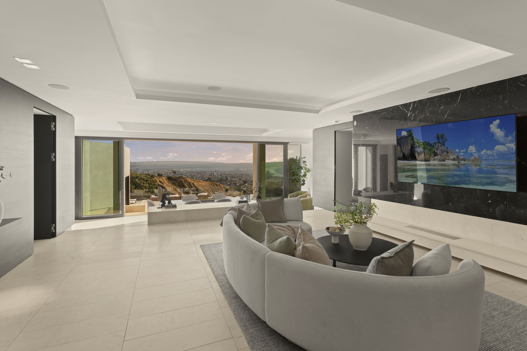 This elegant and contemporary living room features a spacious, open-concept design anchored by a large curved sofa and a sleek black marble fireplace wall with an integrated television. Large floor-to-ceiling glass doors open seamlessly to a generous patio, showcasing breathtaking, panoramic hillside and city views. The neutral color palette, stone flooring, and sophisticated minimalist aesthetic create a luxurious and airy atmosphere suitable for upscale entertaining.
