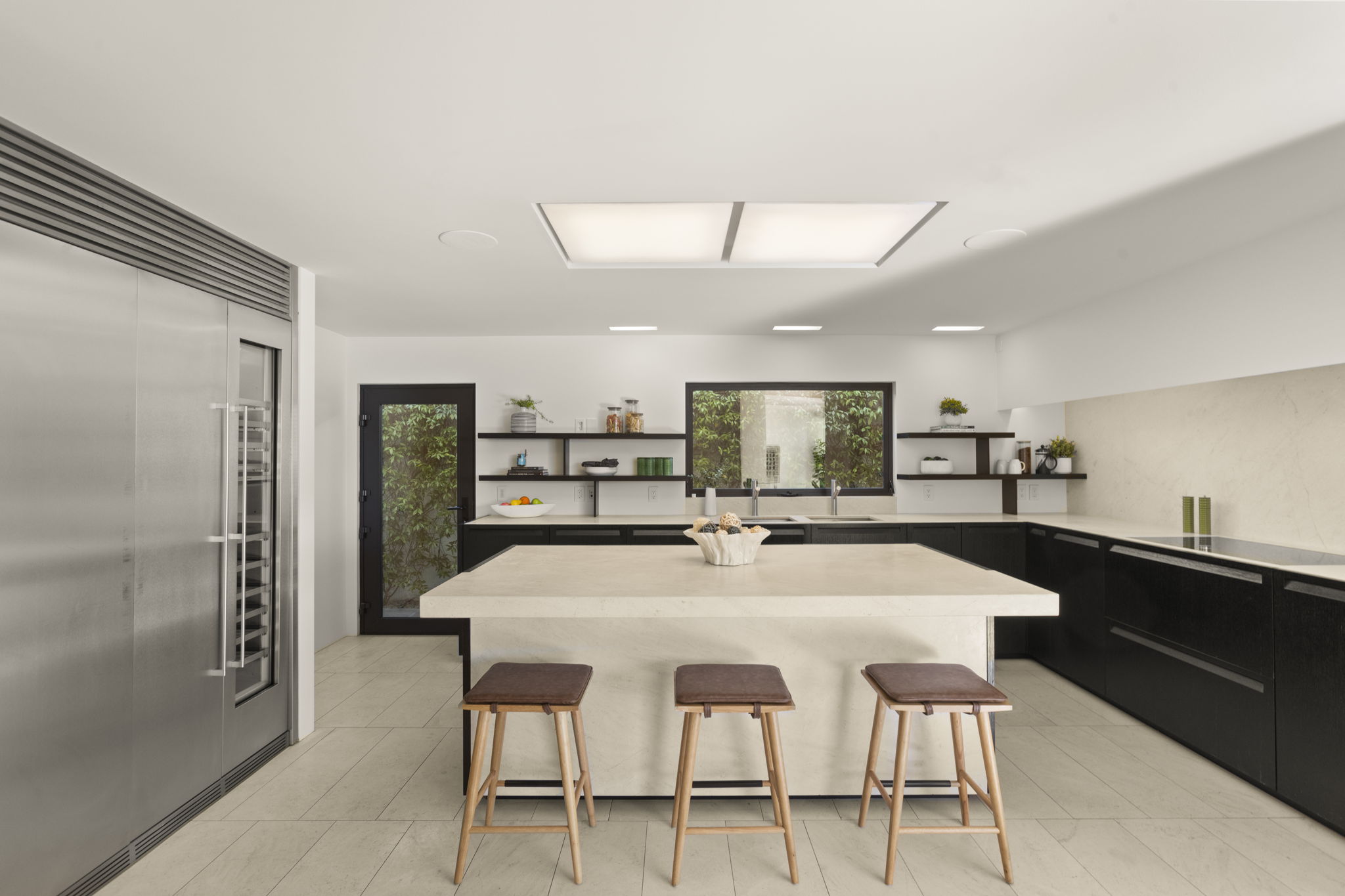 This sleek, modern kitchen features a large central island with light-colored stone countertops and bar stool seating, contrasting with the dark wood cabinetry throughout. The space is well-lit by recessed lighting and a skylight, complemented by integrated stainless steel appliances and a clean, minimalist design. A window over the sink provides a pleasant view of the outdoor greenery, enhancing the bright and airy atmosphere.