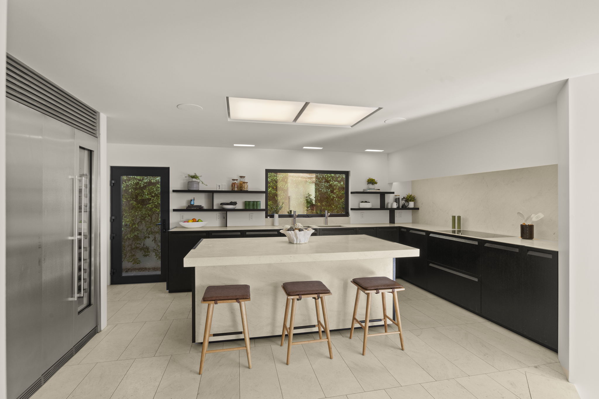 This sleek, modern kitchen features a large central island with quartz countertops and three barstools, set against high-contrast black cabinetry and matching wall accents. The space is anchored by a stainless steel refrigerator and elegant slab-style backsplashes, complemented by floating shelves for added storage. Large, recessed ceiling lights and a window overlooking greenery provide a bright, clean, and contemporary atmosphere ideal for a luxury listing.