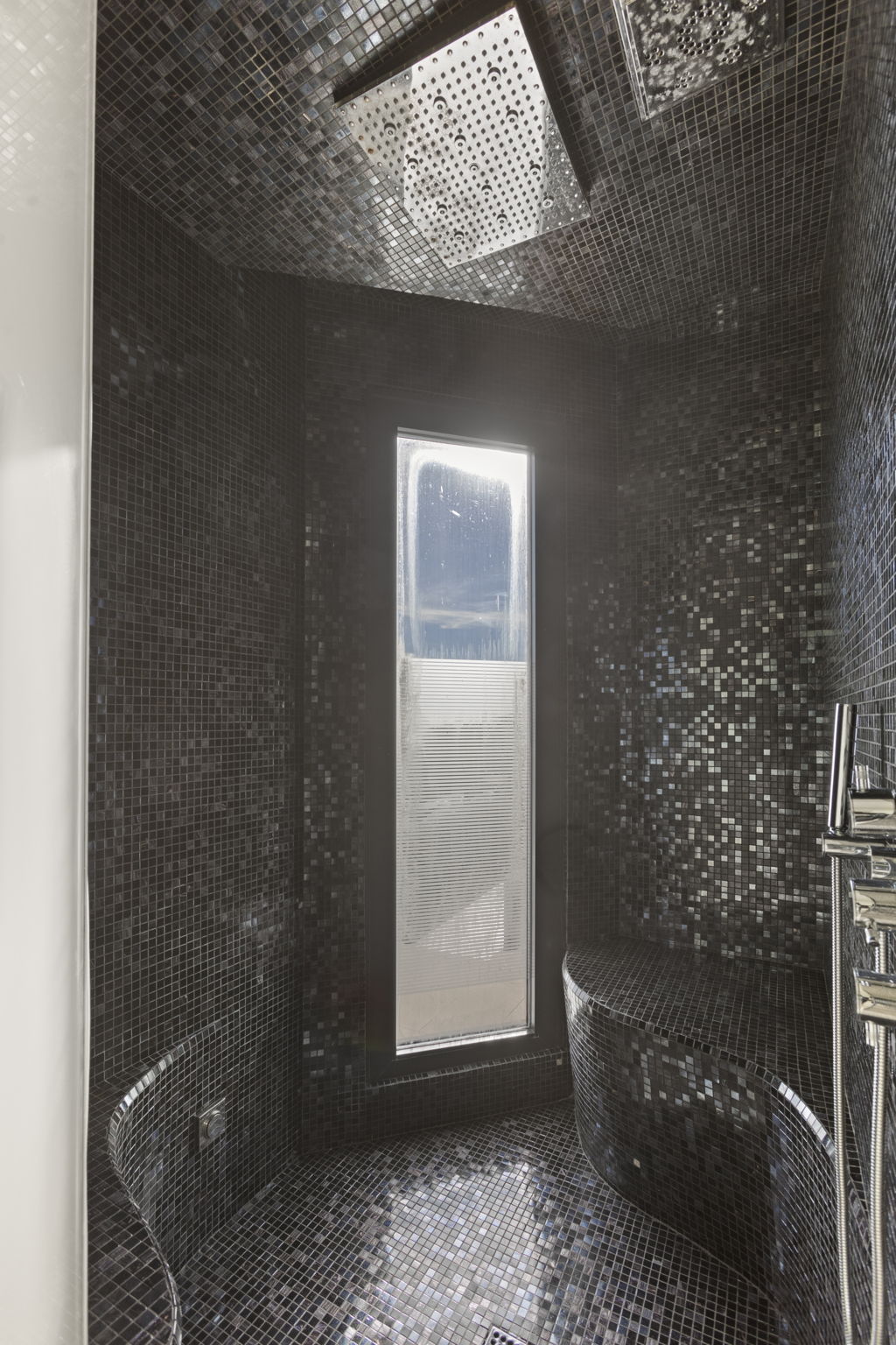 This luxurious, high-end shower features dark, shimmering mosaic tiling that covers the walls, floor, and built-in benches, creating a dramatic and immersive modern aesthetic. It includes a large, professional-grade rainfall showerhead and a sleek handheld body spray, along with a slender vertical window that allows natural light to filter through a frosted privacy panel. The overall design is sophisticated and spa-like, emphasizing premium materials and a moody, contemporary atmosphere.