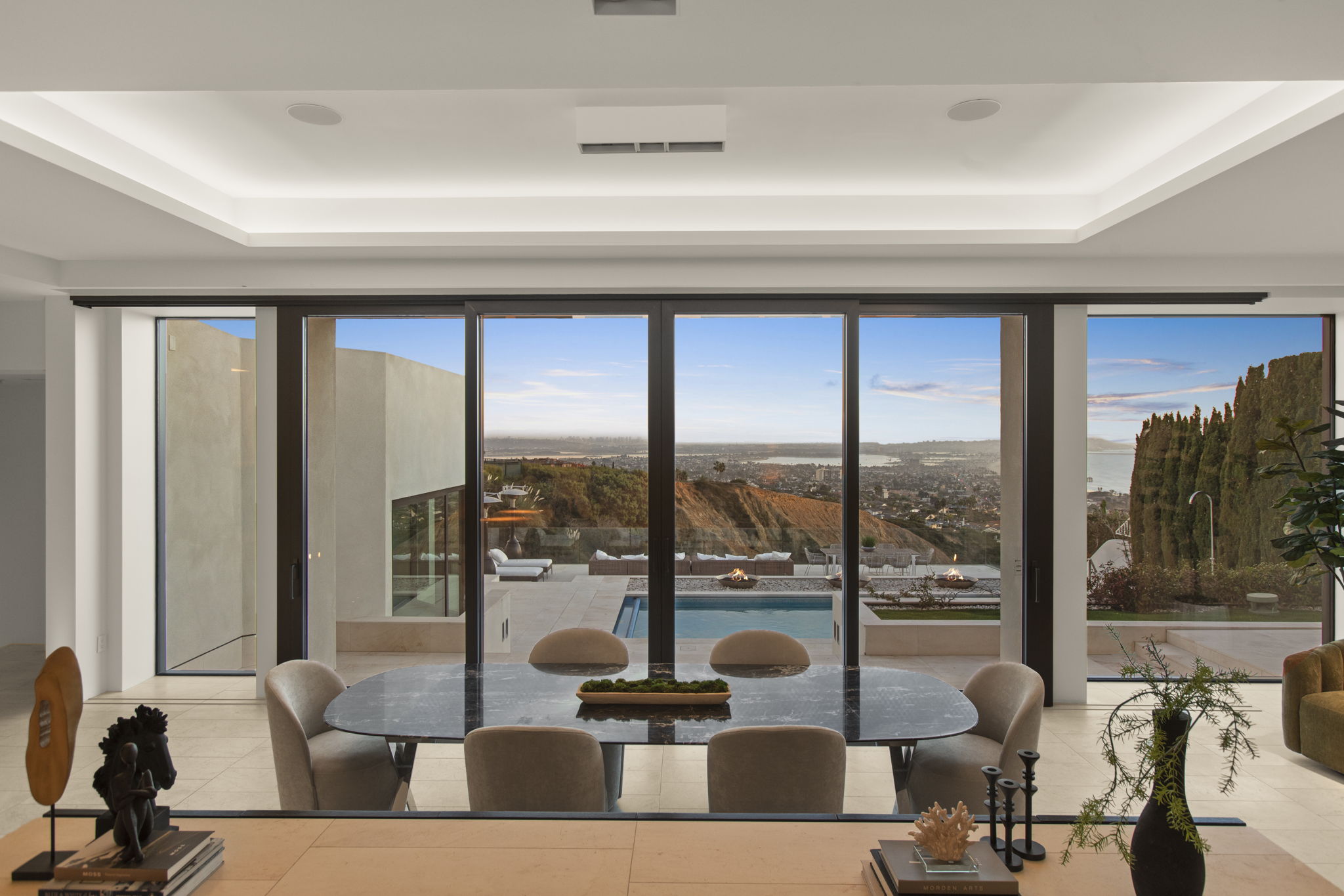 This elegant dining room features a contemporary marble-top table surrounded by plush taupe seating, set against a stunning backdrop of floor-to-ceiling glass sliding doors. The expansive view overlooking a pool, hillside, and city skyline creates a seamless indoor-outdoor living experience. High-end architectural details include recessed cove lighting, a minimalist aesthetic, and sophisticated decor that emphasizes the home's luxurious location.
