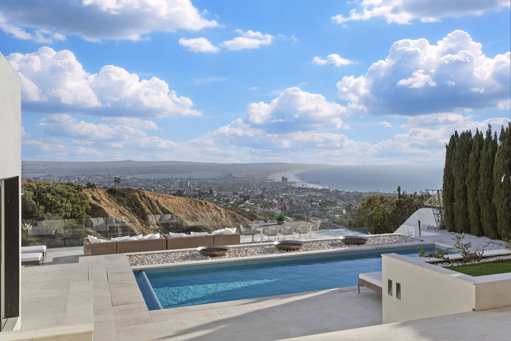 This stunning contemporary outdoor space features a pristine rectangular infinity pool lined with an elegant stone terrace. The area includes comfortable lounge seating and a dining setup, all framing a breathtaking panoramic view of the coastline and ocean horizon. Mature row of cylindrical cypress trees adds architectural interest and privacy to this luxurious cliffside setting.