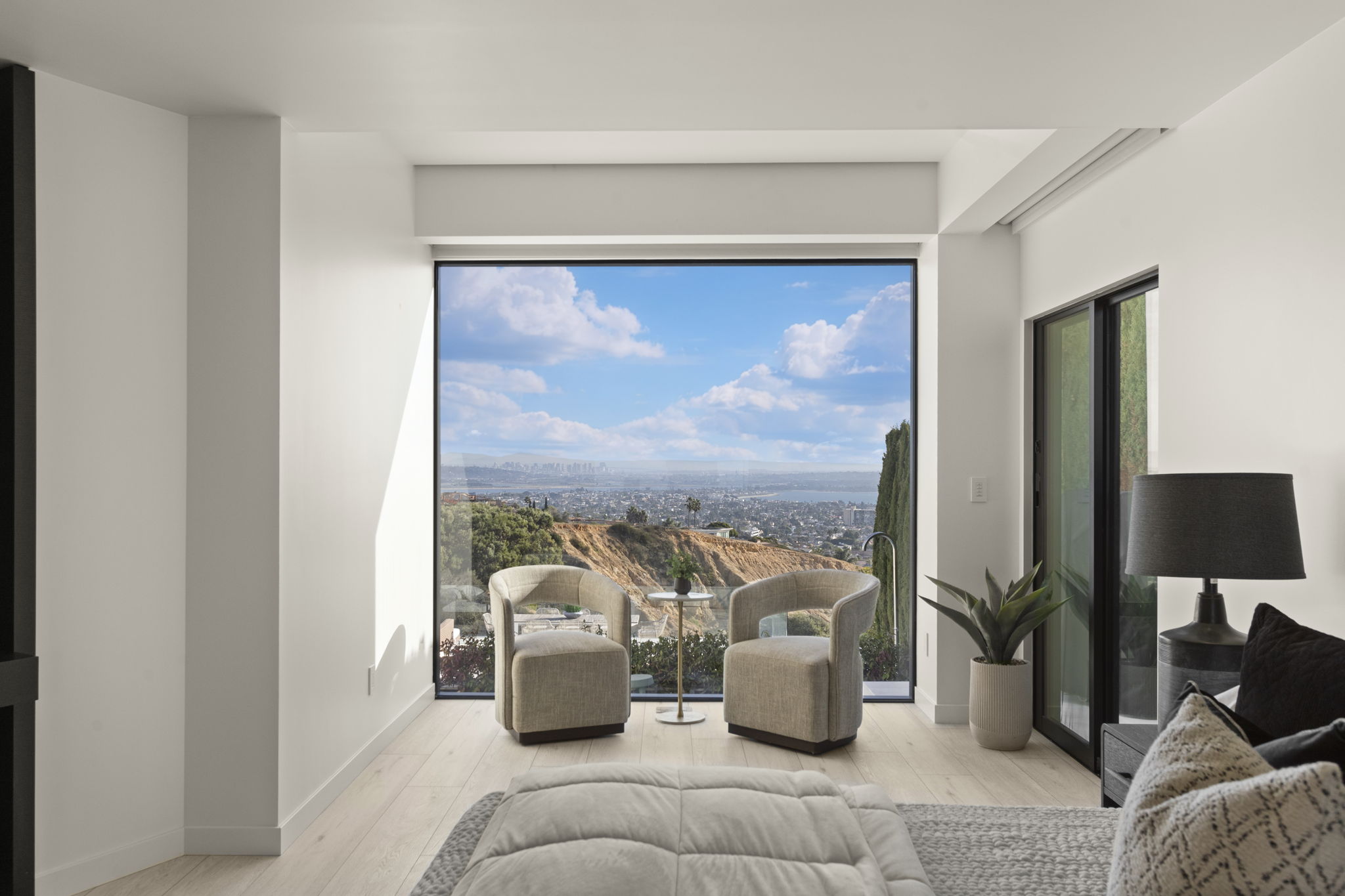 This tranquil primary bedroom features light wood flooring, neutral tone furnishings, and a large floor-to-ceiling picture window that maximizes natural light. The room is anchored by two chic upholstered chairs and a minimalist side table, perfectly positioned to take advantage of the expansive panoramic city and hillside views. The clean, modern aesthetic creates a serene and luxurious retreat space.