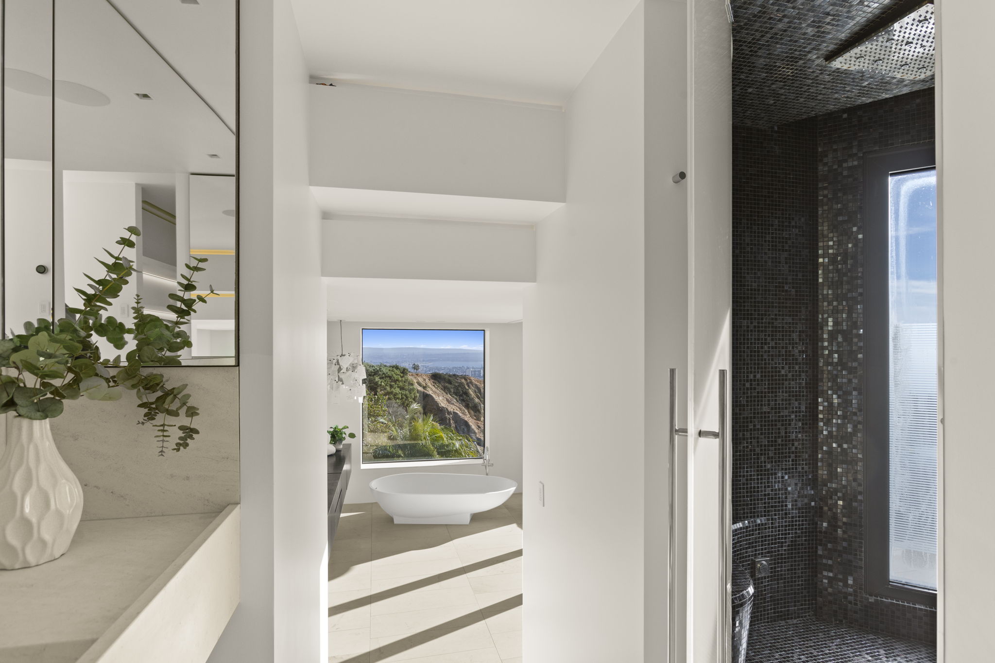 This modern, luxurious primary bathroom features a stunning freestanding soaking tub positioned perfectly in front of a window with sweeping views of the coastline and hills. The space incorporates a sleek, minimalist vanity area to the left and a dramatic, enclosed glass-door shower tiled with dark, shimmering mosaic tiles to the right. The white walls paired with clean lines create an airy, sophisticated atmosphere that emphasizes the home's spectacular location.