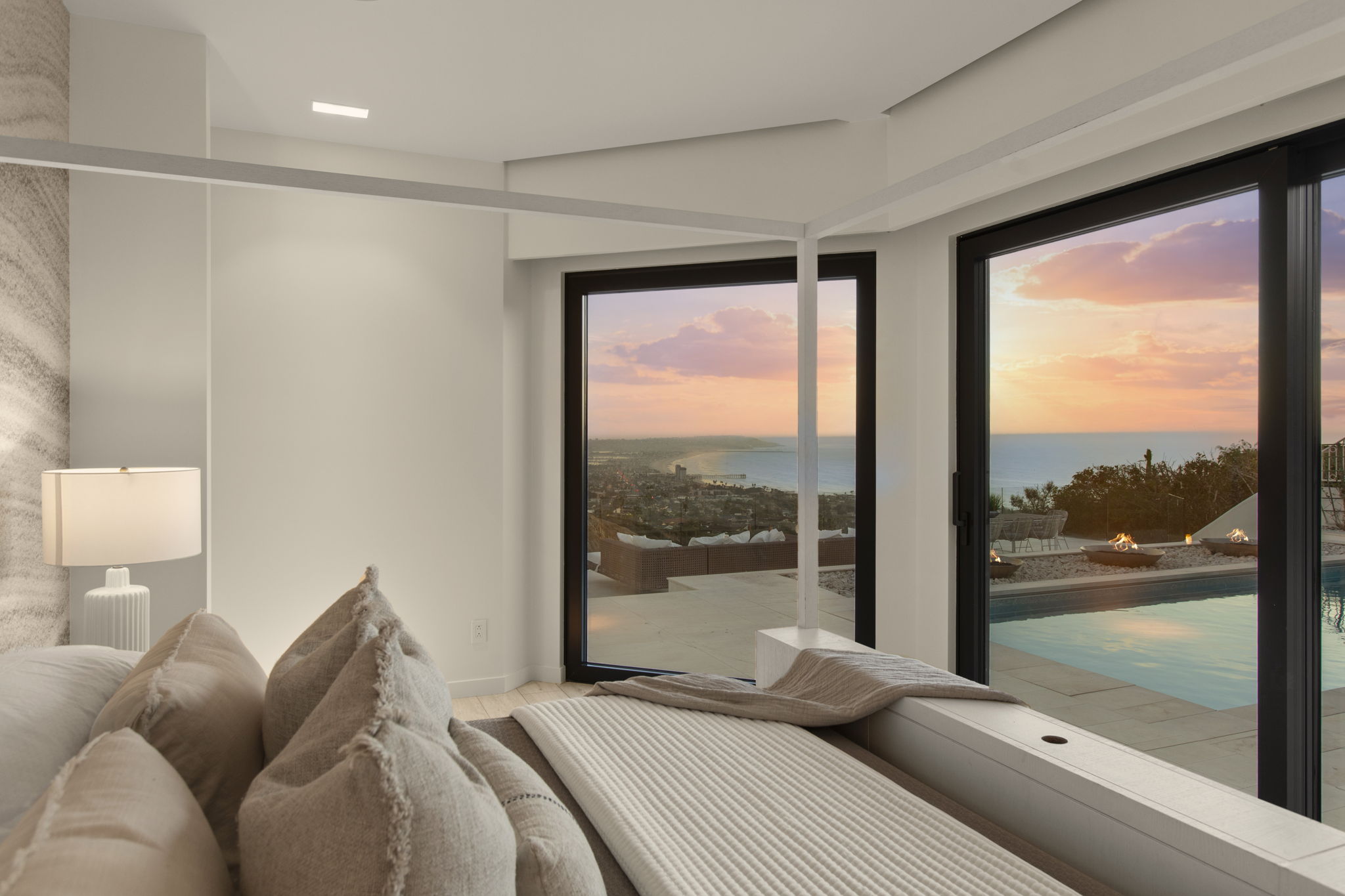This primary bedroom offers a serene, luxurious atmosphere with a contemporary four-poster bed frame and soft, neutral-toned bedding. Floor-to-ceiling glass sliding doors frame a breathtaking panoramic view of the sunset over the coast, seamlessly connecting the interior to a private patio area featuring an infinity pool and fire feature. The space is minimalist and refined, emphasizing the stunning indoor-outdoor flow and high-end coastal lifestyle.
