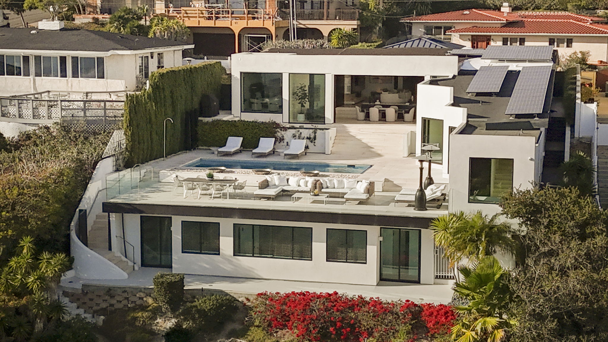 This modern, elevated residence features an expansive multi-level outdoor deck, a sleek swimming pool, and a white architectural aesthetic. The home seamlessly integrates indoor and outdoor living with floor-to-ceiling glass doors and well-appointed seating areas, offering a luxurious and private retreat. Nestled in a lush, terraced landscape with a view of neighboring properties, it presents an impressive and contemporary upscale design.