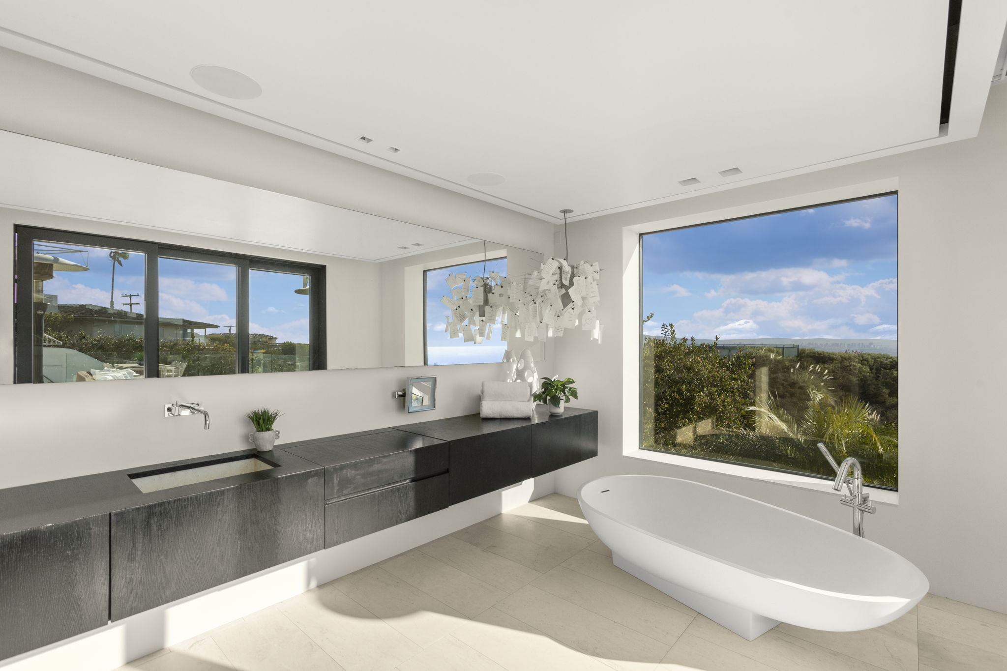 This modern, high-end primary bathroom features a sleek floating vanity with a dark wood finish and an integrated stone sink. A dramatic, organic-shaped sculptural pendant light hangs above the vanity, while a large soaking tub sits by a floor-to-ceiling window offering panoramic views. The space is minimalist and luxurious, combining natural light with contemporary design elements to create a tranquil spa-like atmosphere.