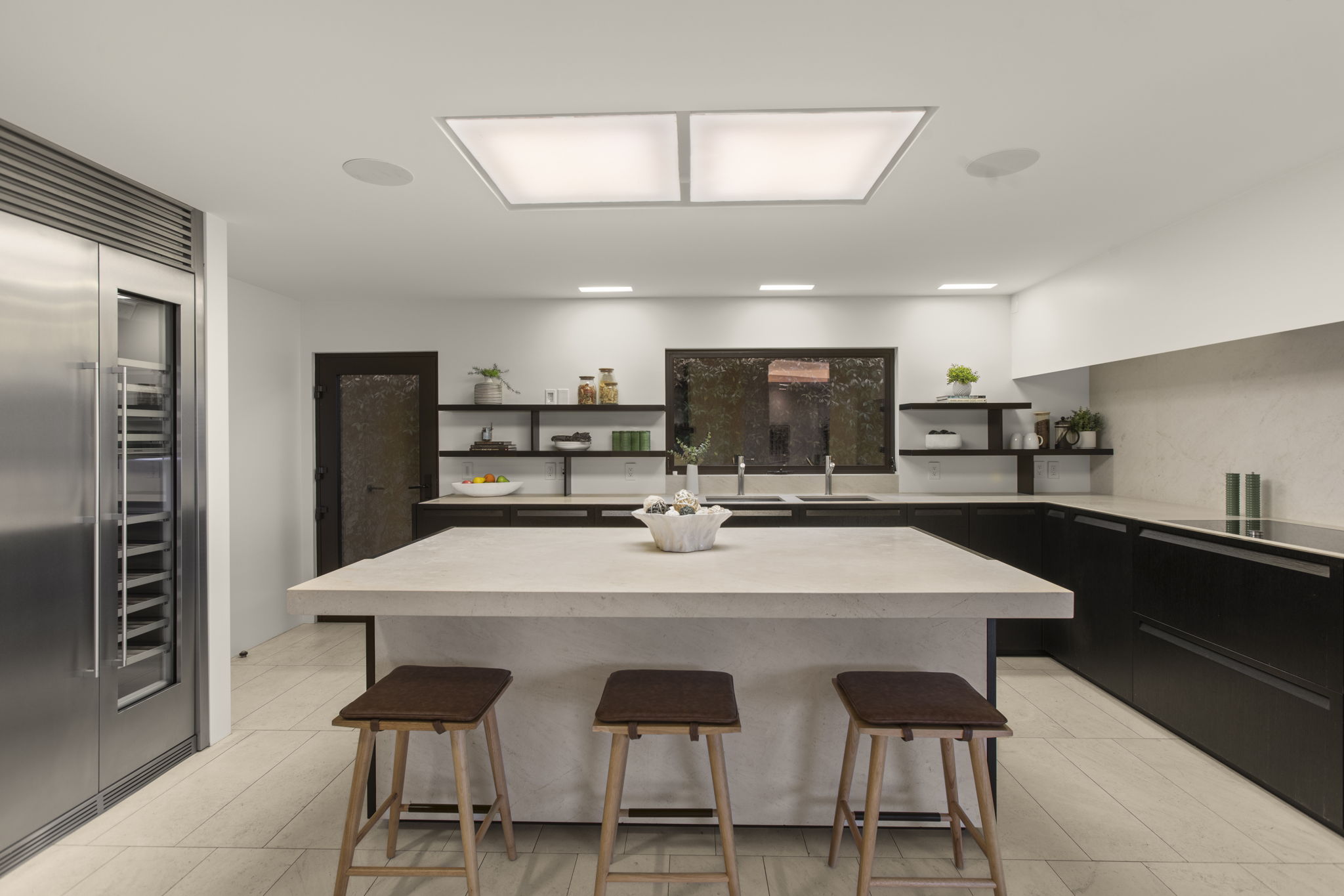 This modern kitchen features dark cabinetry paired with light stone countertops and a matching expansive kitchen island. The space is well-lit by recessed lighting and a large ceiling fixture, showcasing three contemporary bar stools and integrated stainless steel appliances. The clean, minimalist design creates a sleek and sophisticated atmosphere ideal for both cooking and entertaining.