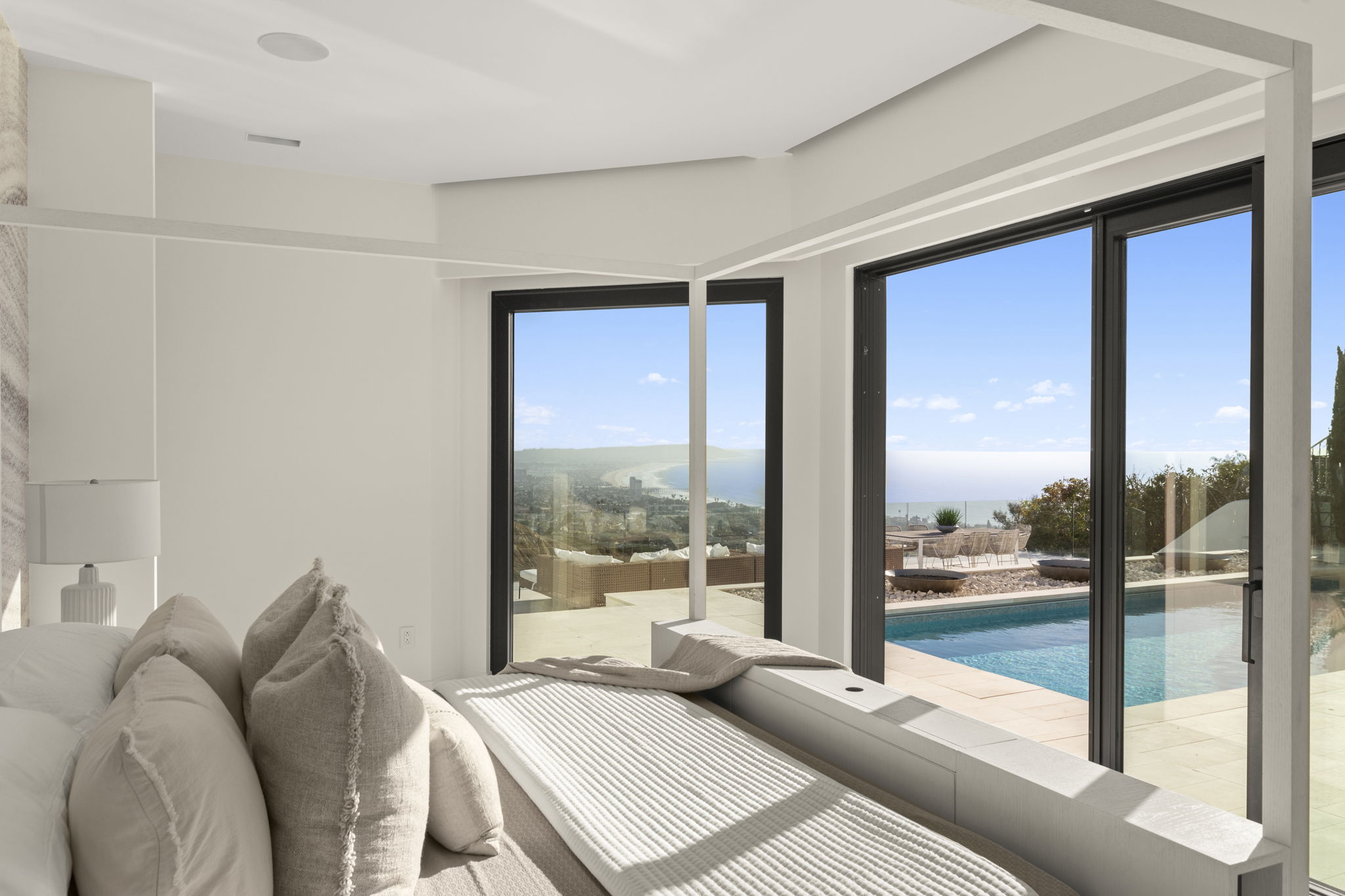 This luxurious primary bedroom features a modern, minimalist design with a clean white palette and a prominent canopy bed frame. The room offers stunning, expansive floor-to-ceiling glass sliding doors that seamlessly blend indoor living with an outdoor terrace, private pool, and breathtaking panoramic ocean views. The tranquil, light-filled space emphasizes a sophisticated indoor-outdoor lifestyle, perfect for capturing scenic coastal scenery.