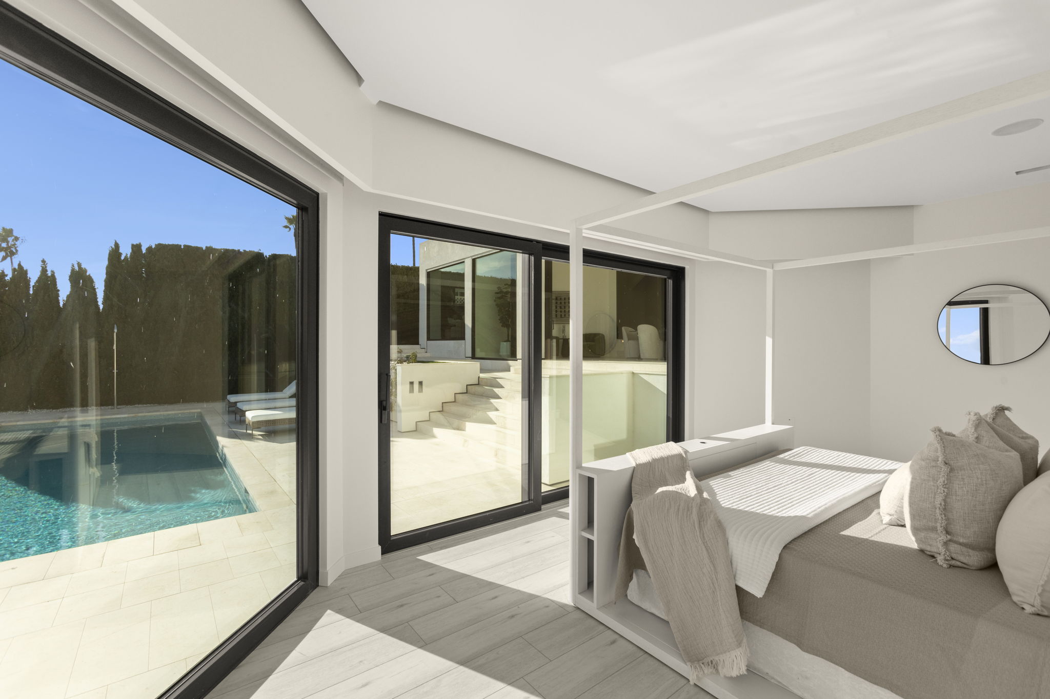 This tranquil primary bedroom features a modern canopy bed and floor-to-ceiling sliding glass doors that seamlessly connect the indoor living space to an outdoor pool and patio area. The light-filled room is decorated in a sophisticated neutral palette, characterized by minimalist decor, light wood flooring, and a serene atmosphere. The presence of the sparkling pool and clean architectural lines enhances the luxurious, resort-like feel of this home.