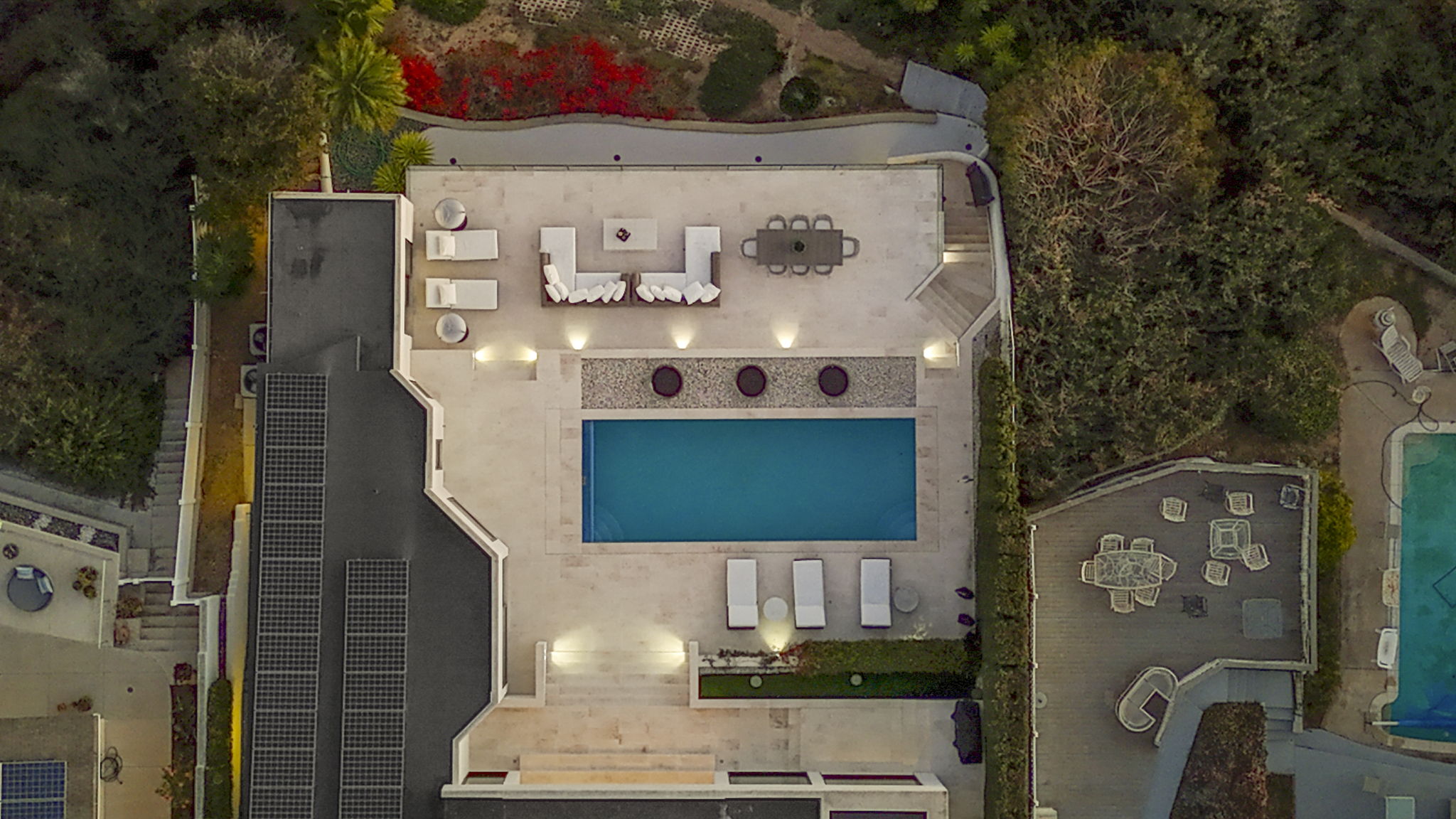 This high-angle, top-down aerial view captures an expansive, luxurious outdoor living area centered around a rectangular swimming pool. The patio features multiple seating arrangements, modern loungers, and warm accent lighting, creating a perfect space for entertainment and relaxation. The surrounding lush landscaping and transition to an adjacent wooden deck enhance the property's elegant and private atmosphere.
