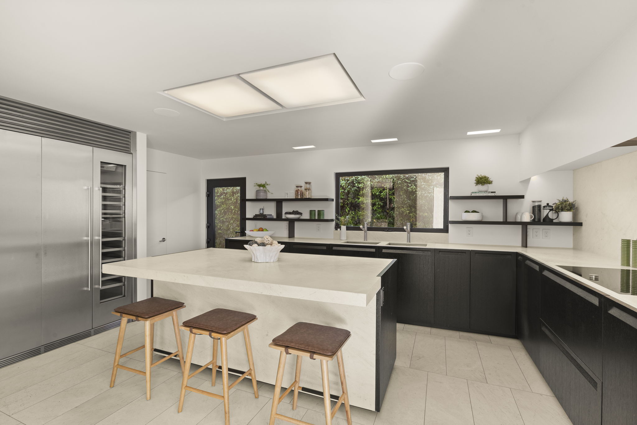This sleek, contemporary kitchen features high-contrast dark wood cabinetry paired with light stone countertops and a spacious central island. The room is accented by minimalist open shelving, professional-grade stainless steel appliances, and a seamless flow that creates a clean, sophisticated aesthetic. Large, recessed lighting fixtures and a window overlooking greenery complete this modern culinary space.