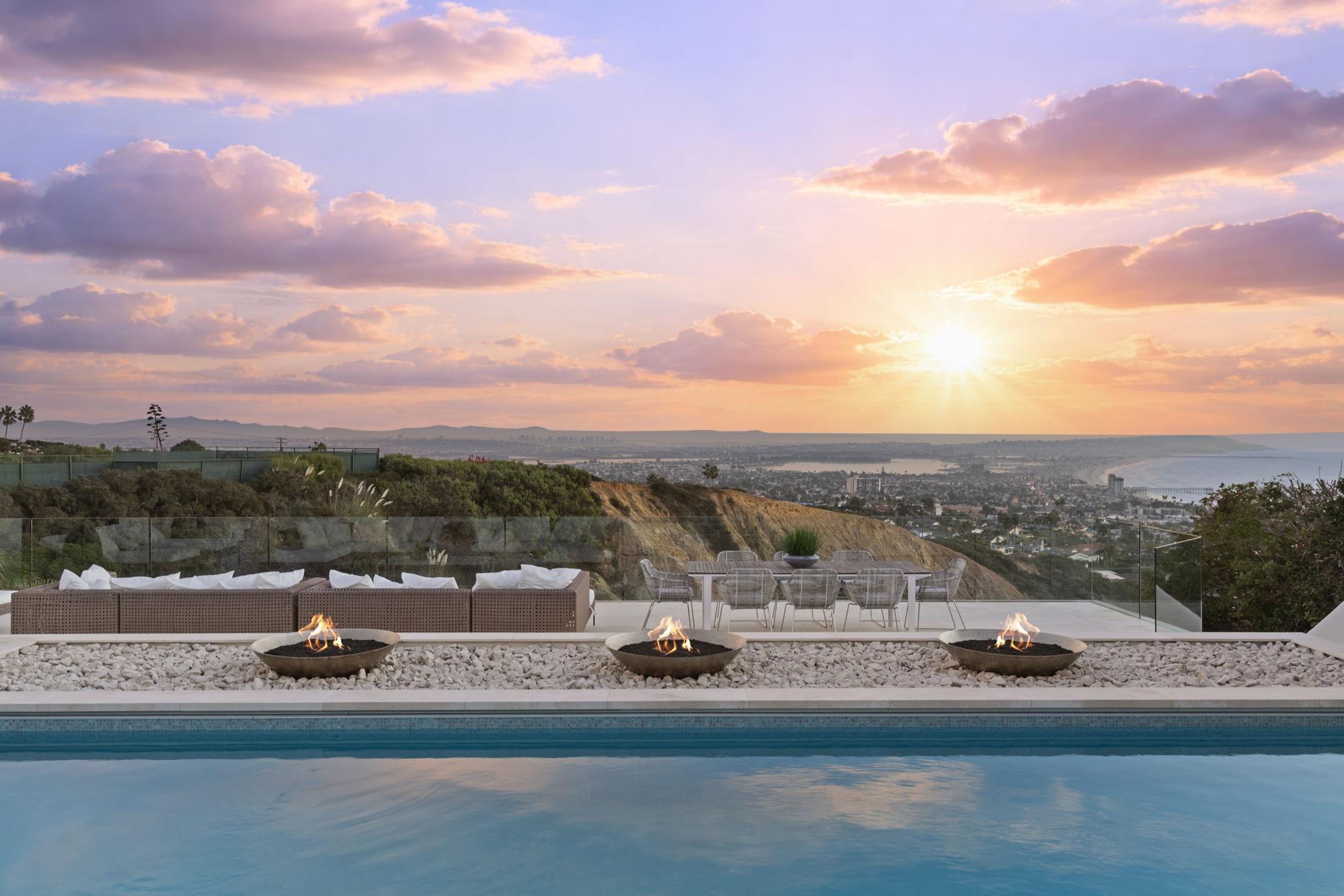 This stunning outdoor space features a serene infinity-edge pool bordered by three modern fire bowls and a rocky garden bed. A spacious patio area includes a comfortable, modern outdoor seating arrangement and a dining set, all framed by a glass railing that offers breathtaking panoramic sunset views over the coastal city below. The atmosphere is one of sophisticated luxury, perfectly curated for high-end outdoor entertaining.