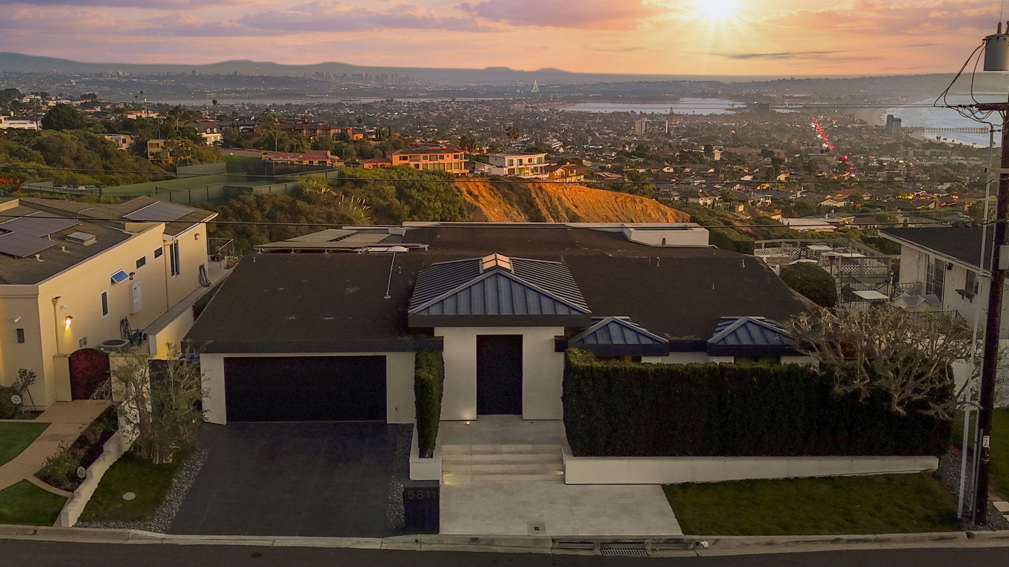 This stunning contemporary residence is captured from an elevated perspective, showcasing its clean lines and prominent blue metal roofing accents. The home features a balanced facade with a private gated entry and driveway, situated prominently to capture the breathtaking sunset views over the coastal city below. Its prime location combines architectural elegance with a spectacular panoramic backdrop, making it a compelling property for those seeking both style and scenery.