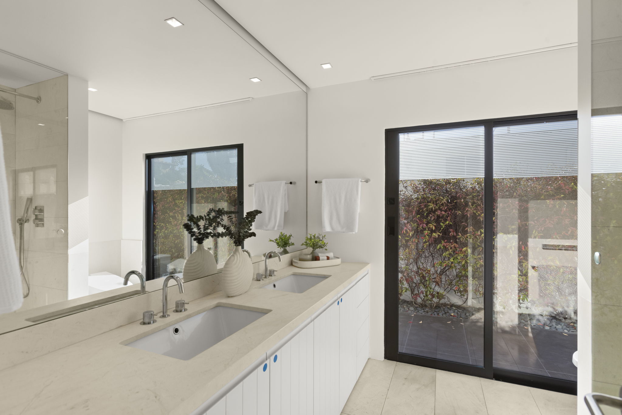This modern primary bathroom features a sleek double vanity with quartz countertops, integrated white sinks, and contemporary chrome fixtures. A large floor-to-ceiling mirror enhances the sense of space, while expansive sliding glass doors provide a seamless connection to a private, sunlit outdoor garden area. The minimalist design, accented by warm neutral tones and subtle greenery, creates a tranquil and sophisticated atmosphere.