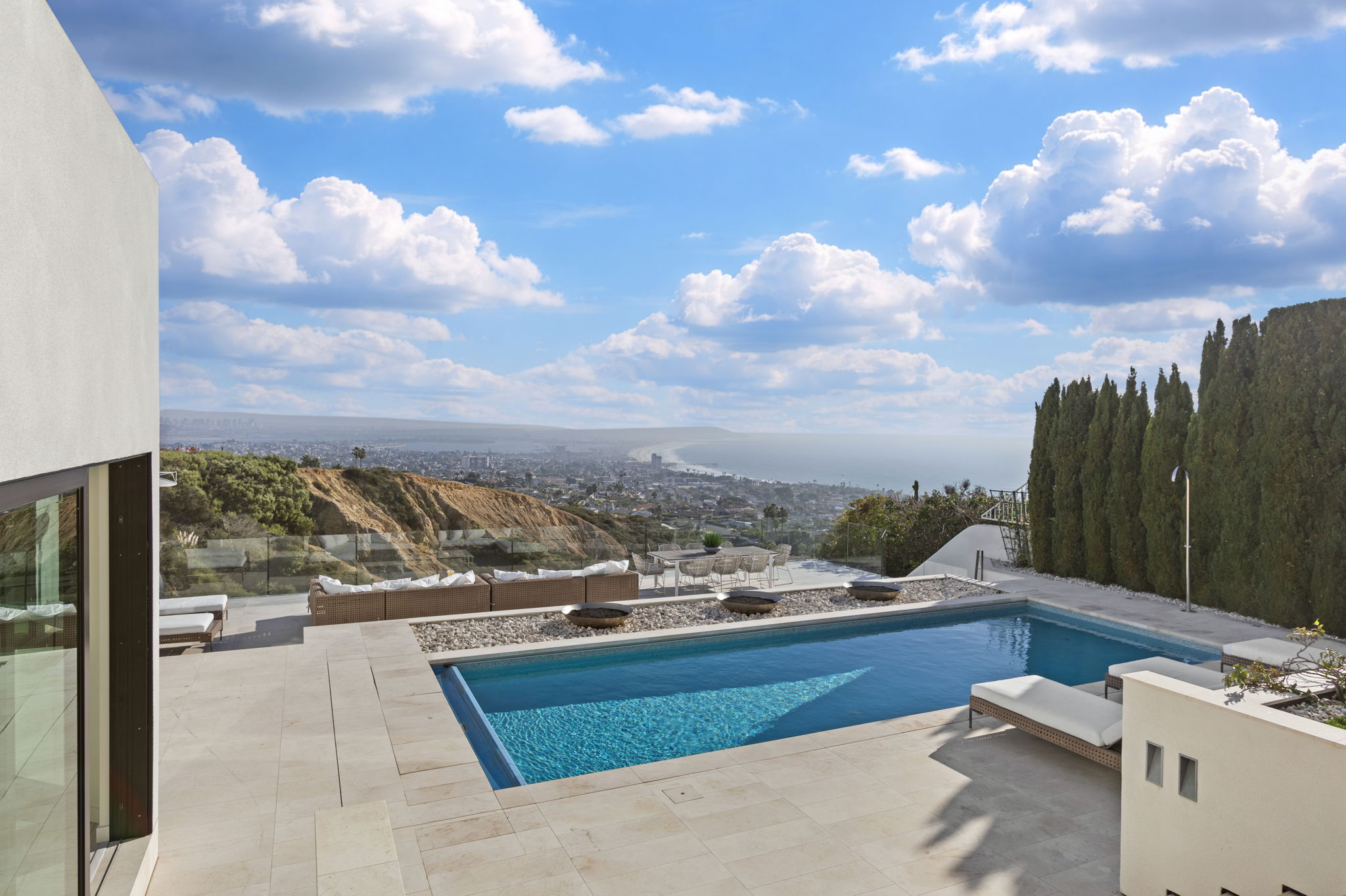 This stunning outdoor space features a rectangular swimming pool set against a breathtaking panoramic view of the coastline and ocean. The pool deck is finished in large, light-colored stone pavers and includes lounge chairs, an outdoor seating area, and fire pits, creating an ideal setting for luxury entertaining. The property boasts a contemporary design that seamlessly integrates indoor-outdoor living with a cliffside backdrop.