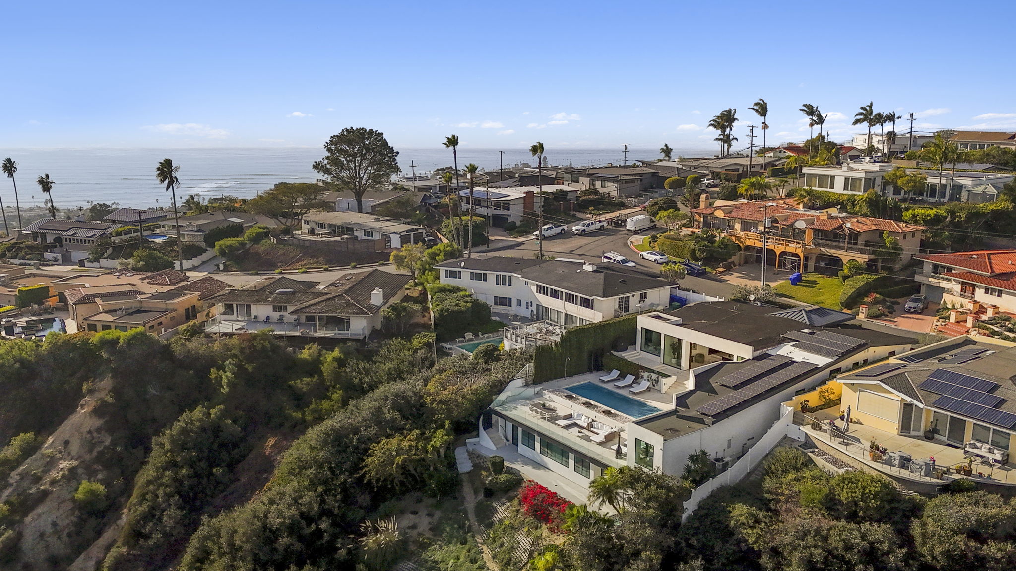 This stunning high-angle aerial view captures a coastal residential neighborhood, showcasing contemporary homes with prime ocean views. The featured property in the foreground boasts multiple levels, an inviting infinity pool, rooftop solar panels, and spacious terraces designed for indoor-outdoor living. The lush landscaping and proximity to the Pacific Ocean create a highly desirable and luxurious setting.