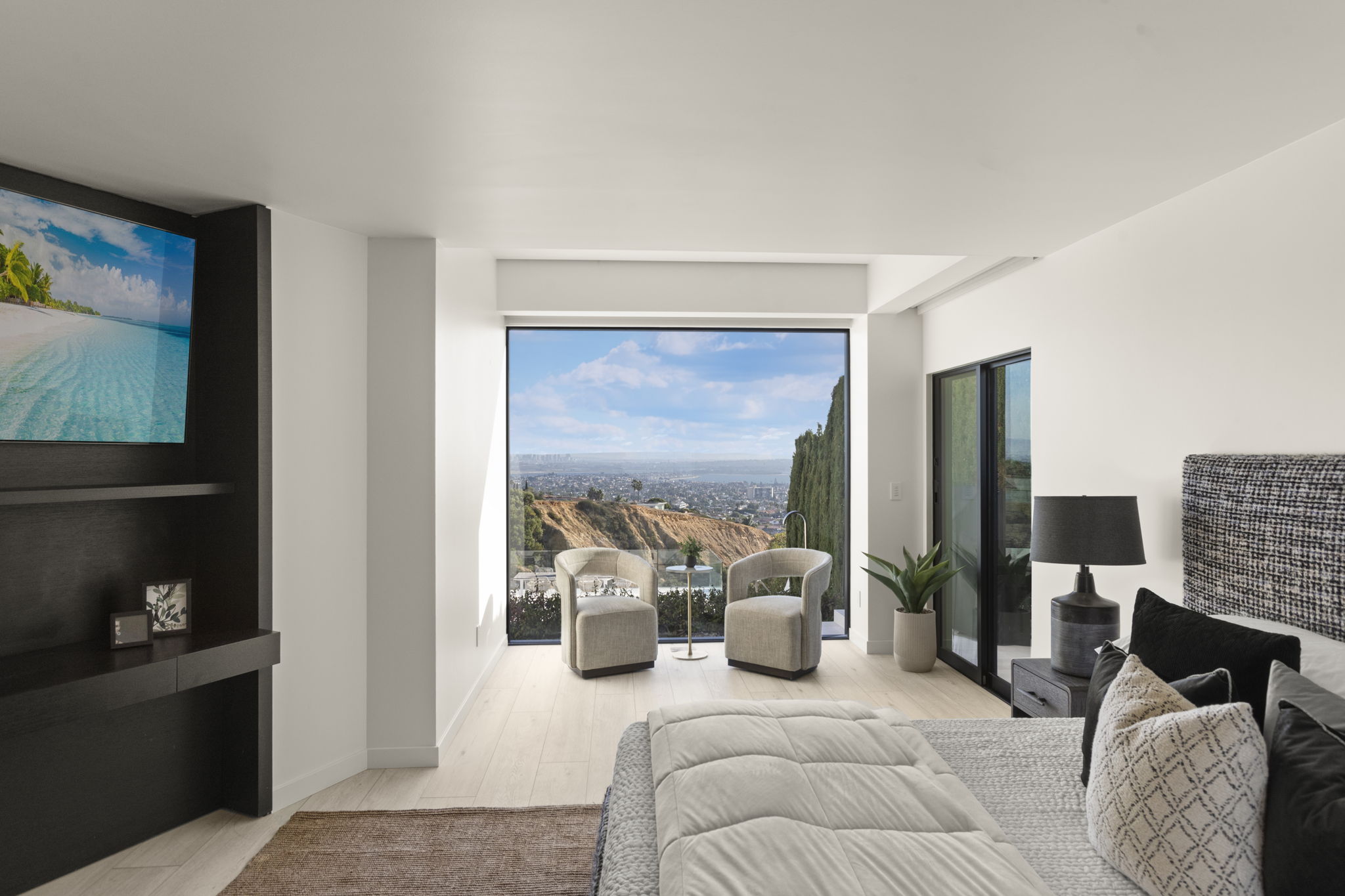 This modern, spacious primary bedroom features a clean, minimalist aesthetic with neutral tones and high-end finishes. A large floor-to-ceiling window serves as the focal point, offering panoramic sunset views, while a built-in media console and well-appointed furniture create a luxurious retreat. The room is seamlessly connected to the outdoors through sliding glass doors and provides an serene, elevated living experience.