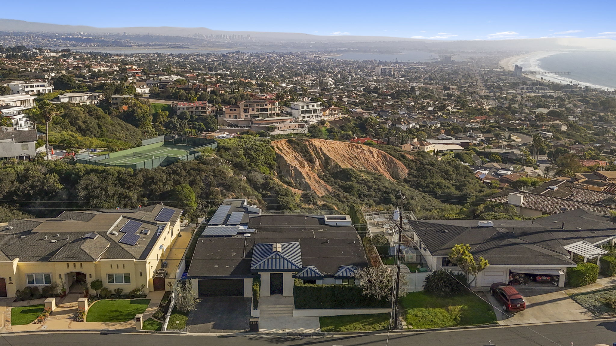 This aerial view captures a contemporary residential home situated prominently in a hilly suburban neighborhood with a scenic coastal overlook. The property features solar panels on its flat roofing, a clean architectural design with distinct blue striped accents, and an attached driveway. The setting offers expansive panoramic views of the city valley, surrounding lush green terrain, and the distant ocean coastline, making it a highly desirable location.
