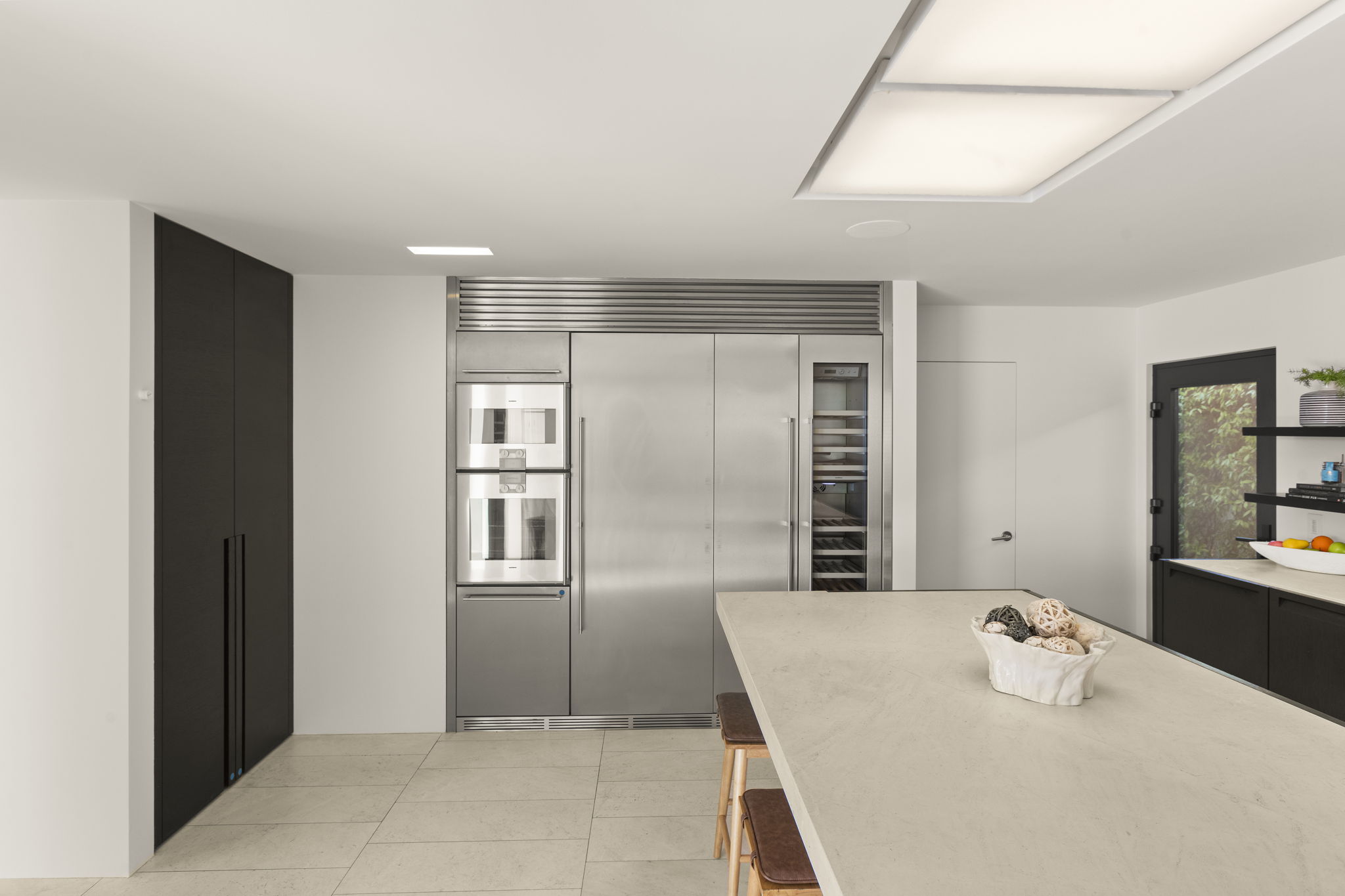 This sleek, modern kitchen features high-end stainless steel appliances, including an integrated refrigerator and wall ovens, contrasted by minimalist dark cabinetry. A large, clean-lined island with a textured stone countertop serves as a central focal point, complemented by subtle under-cabinet lighting and recessed ceiling lights. The space offers a sophisticated, contemporary aesthetic with a neutral palette and clean transitions between textures.