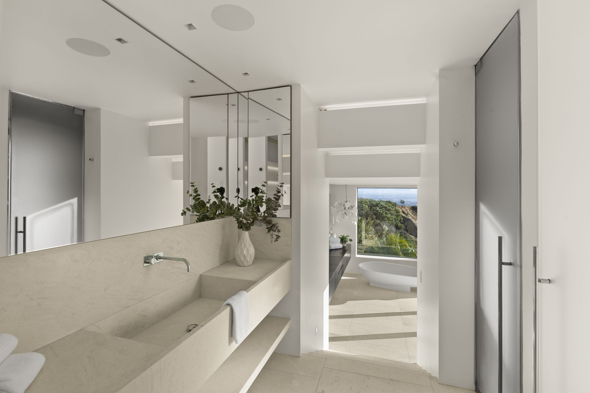 This luxurious primary bathroom features a minimalist, contemporary aesthetic with a custom stone trough sink and a large vanity mirror. A seamless transition leads into an adjacent bathing area with a soaking tub positioned in front of a floor-to-ceiling window showcasing expansive, scenic views. The space is characterized by its light, neutral palette and sophisticated, modern design.