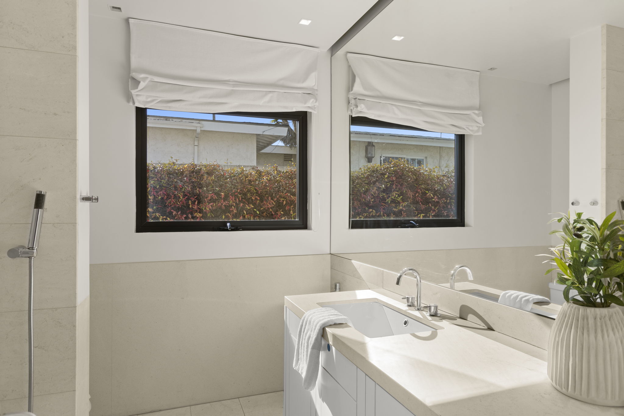 This modern bathroom features a sleek vanity with a light-colored countertop, a white under-mount sink, and contemporary chrome fixtures. A large mirror and window with a clean white roman shade help brighten the space, while a textured plant adds a decorative touch. The neutral color palette and minimalist design create a serene and polished environment.