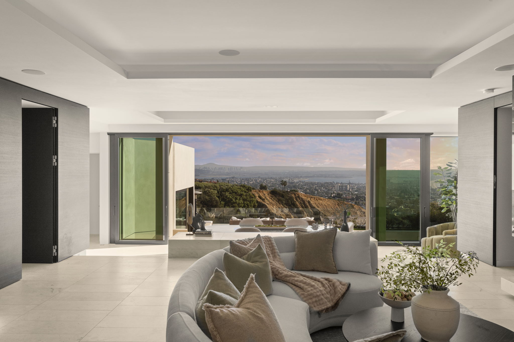 This sophisticated, contemporary living room features a dramatic open-concept layout with expansive pocket doors that seamlessly blend the interior with a breathtaking panoramic coastal view. A sleek, curved sectional sofa centers the room, creating an inviting space that highlights the home's high-end finishes and seamless indoor-outdoor transition.