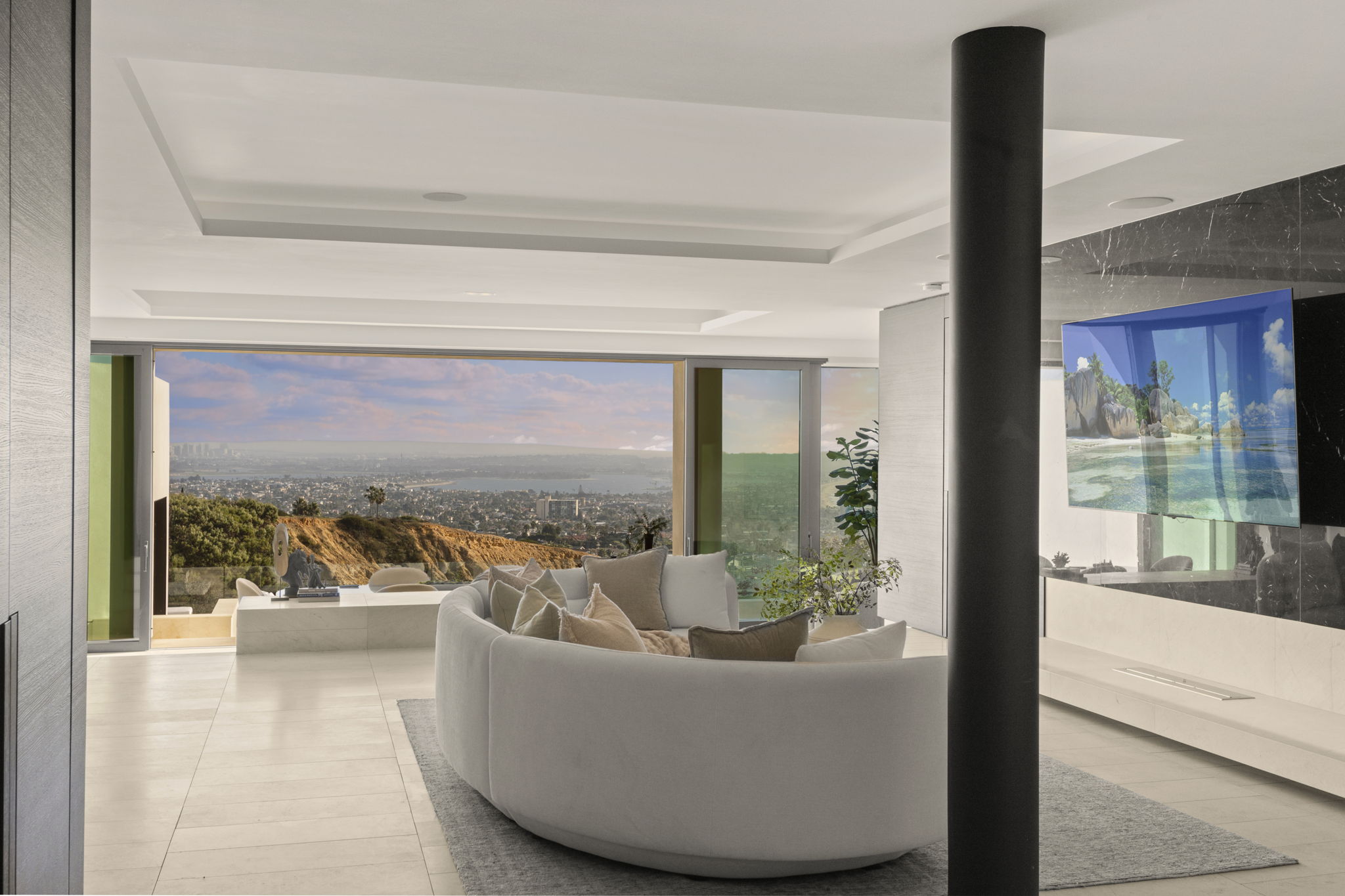 This sophisticated, contemporary living room features an expansive, curved sectional sofa positioned to maximize the breathtaking, wide-angle panoramic view of the coastline and city below. The space is anchored by a striking black cylindrical support pillar and a large television mounted on a sleek, dark-veined marble feature wall. With its neutral color palette, clean lines, and seamless transition to the outdoors through floor-to-ceiling glass doors, the room exudes luxury and a sense of open-concept tranquility.