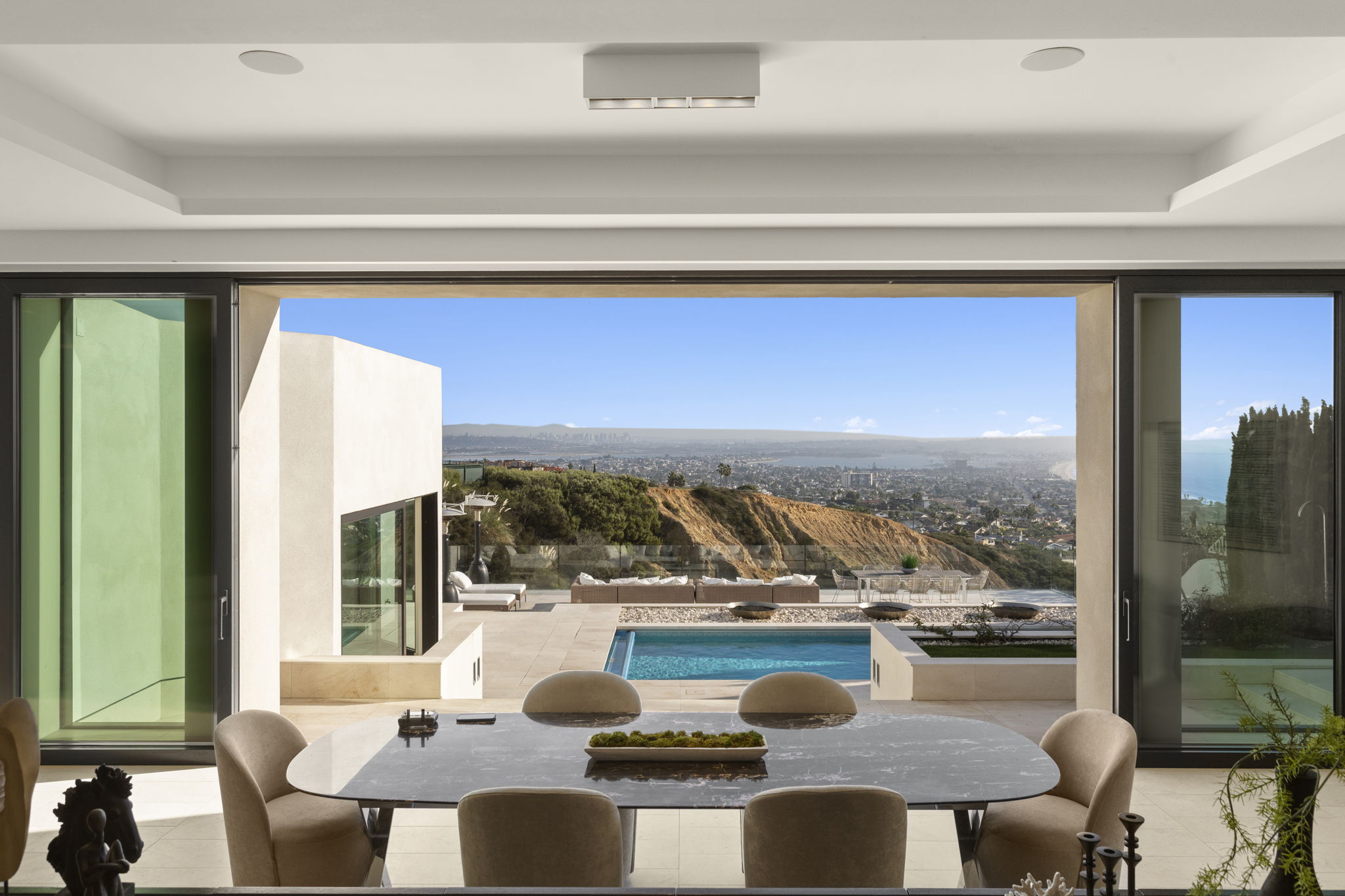 This elegant dining room features a modern, oval marble-topped table surrounded by comfortable taupe upholstered chairs, creating an inviting space for gatherings. The room seamlessly transitions to the outdoors through expansive sliding floor-to-ceiling glass doors that reveal a stunning infinity pool, a coastal cliffside terrace, and panoramic ocean and city views. The transition between the sleek indoor aesthetic and the breathtaking natural vista creates a luxurious and airy atmosphere characteristic of high-end coastal design.