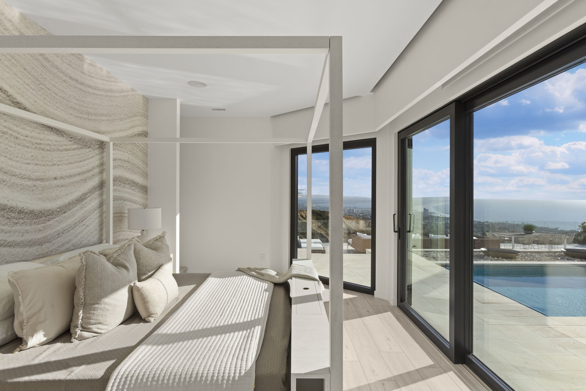 This luxurious primary bedroom features a modern four-poster bed as its centerpiece, set against a stunning custom textured accent wall. The room boasts floor-to-ceiling glass sliding doors that provide seamless access to a terrace and highlight an expansive, panoramic coastal view including a private swimming pool. The interior design emphasizes clean, minimalist aesthetics with a soft, neutral color palette that complements the bright, sun-drenched atmosphere.