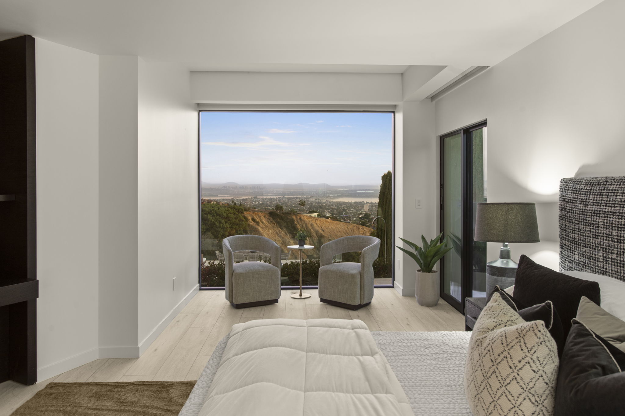 This elegantly appointed primary bedroom features light wood-look flooring and a refined, contemporary aesthetic. A standout feature is the large floor-to-ceiling picture window providing expansive, scenic views of the coastal city and landscape, complemented by a cozy seating area for two. The room is anchored by a high-quality, textured bed ensemble, creating a serene and luxurious retreat.
