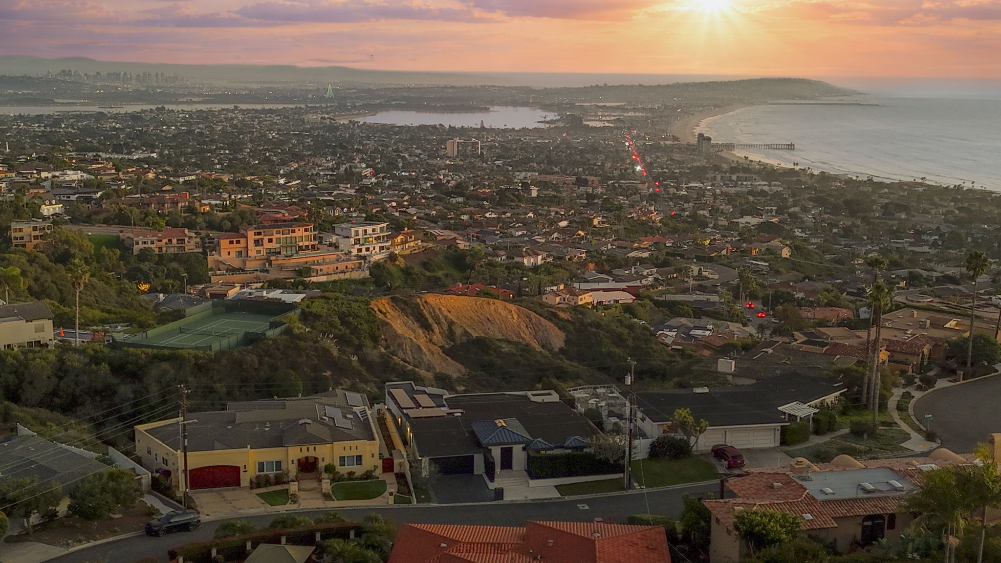 This is a sweeping aerial view of a coastal neighborhood during sunset, showcasing the proximity of hillside estates to the beach and a nearby bay. The scene highlights residential properties with varied Mediterranean-style architecture, lush landscaping, and a panoramic vantage point of the Pacific coastline. The dramatic sky and urban density create a prestigious and desirable setting.
