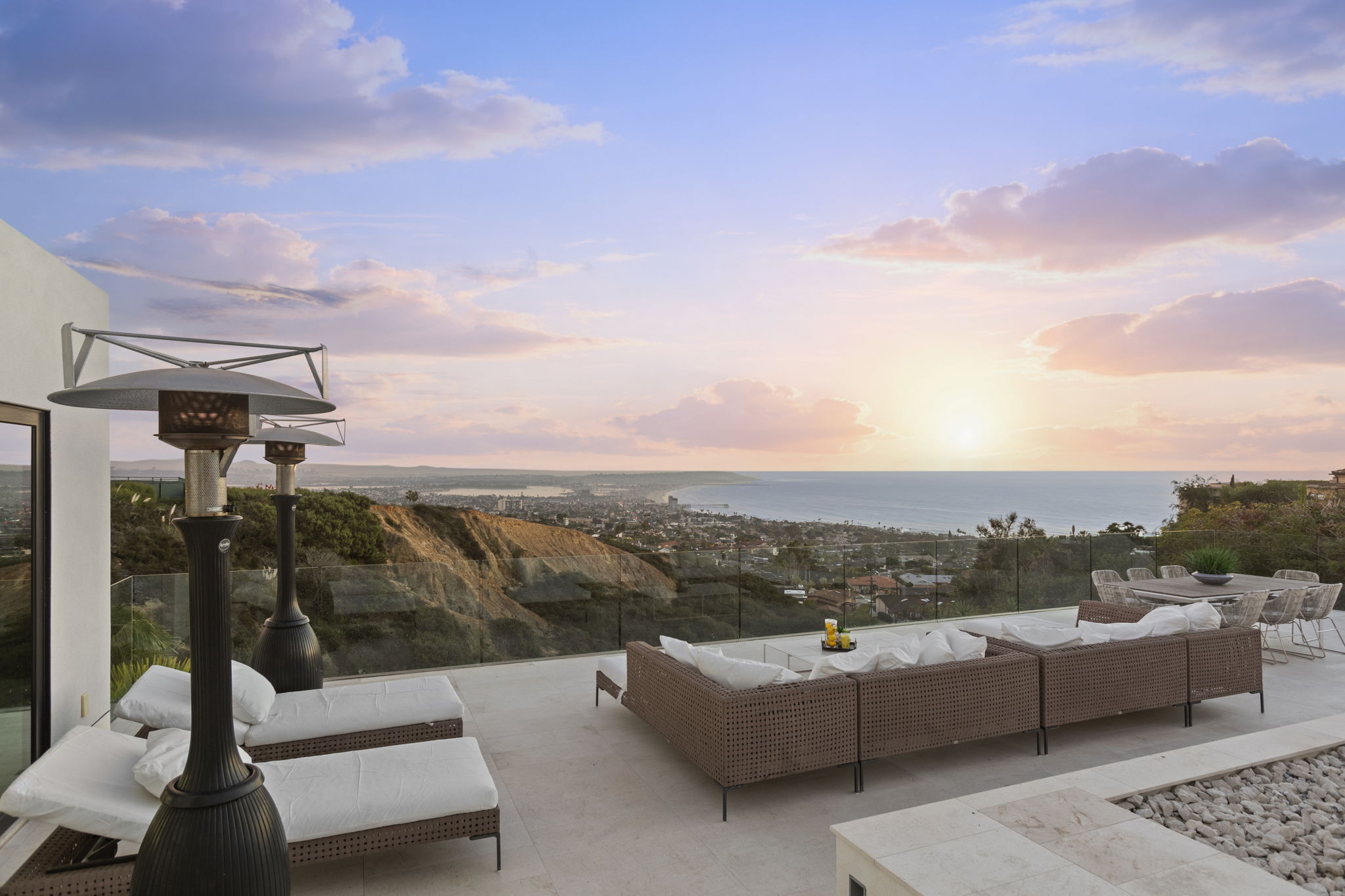 This spacious, modern patio offers a breathtaking panoramic view of the Pacific coast and the residential skyline during a scenic sunset. The outdoor living area is well-equipped with comfortable wicker sofa sectional seating, lounge chairs, and a dining area, all protected by a sleek glass railing. Dual outdoor heaters ensure the space is perfect for evening entertaining, creating a luxurious and serene atmosphere.