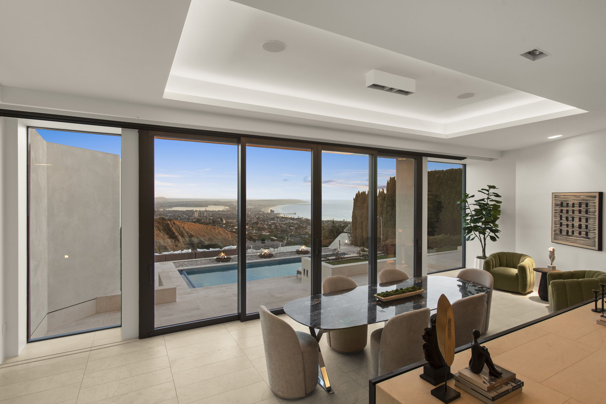 This modern dining area features an elegant oval-shaped marble-topped table surrounded by sleek, beige upholstered chairs. Large, floor-to-ceiling glass sliding doors provide a seamless transition to a private pool deck and offer a breathtaking panoramic view of the coastline and ocean. The space is tastefully decorated with contemporary accents, including green accent chairs and artistic decor, creating a sophisticated and airy atmosphere.