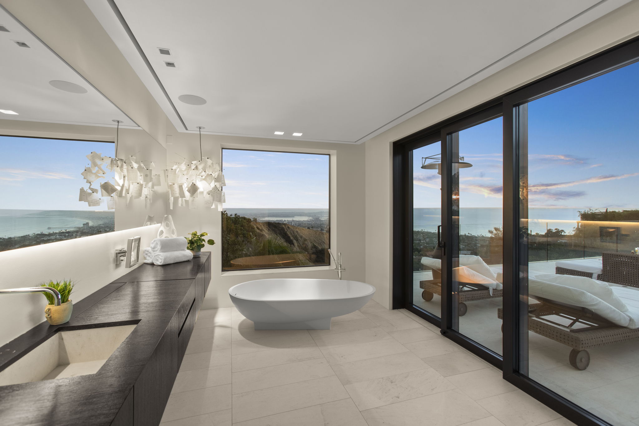 This luxurious primary bathroom features a sleek, minimalist aesthetic with a freestanding white soaking tub as the centerpiece. A floating dark wood vanity runs along the wall, complemented by a large sculptural chandelier and a stunning floor-to-ceiling view of the ocean coast. Glass sliding doors open directly onto a private balcony, creating a seamless indoor-outdoor living experience with a serene, modern vibe.