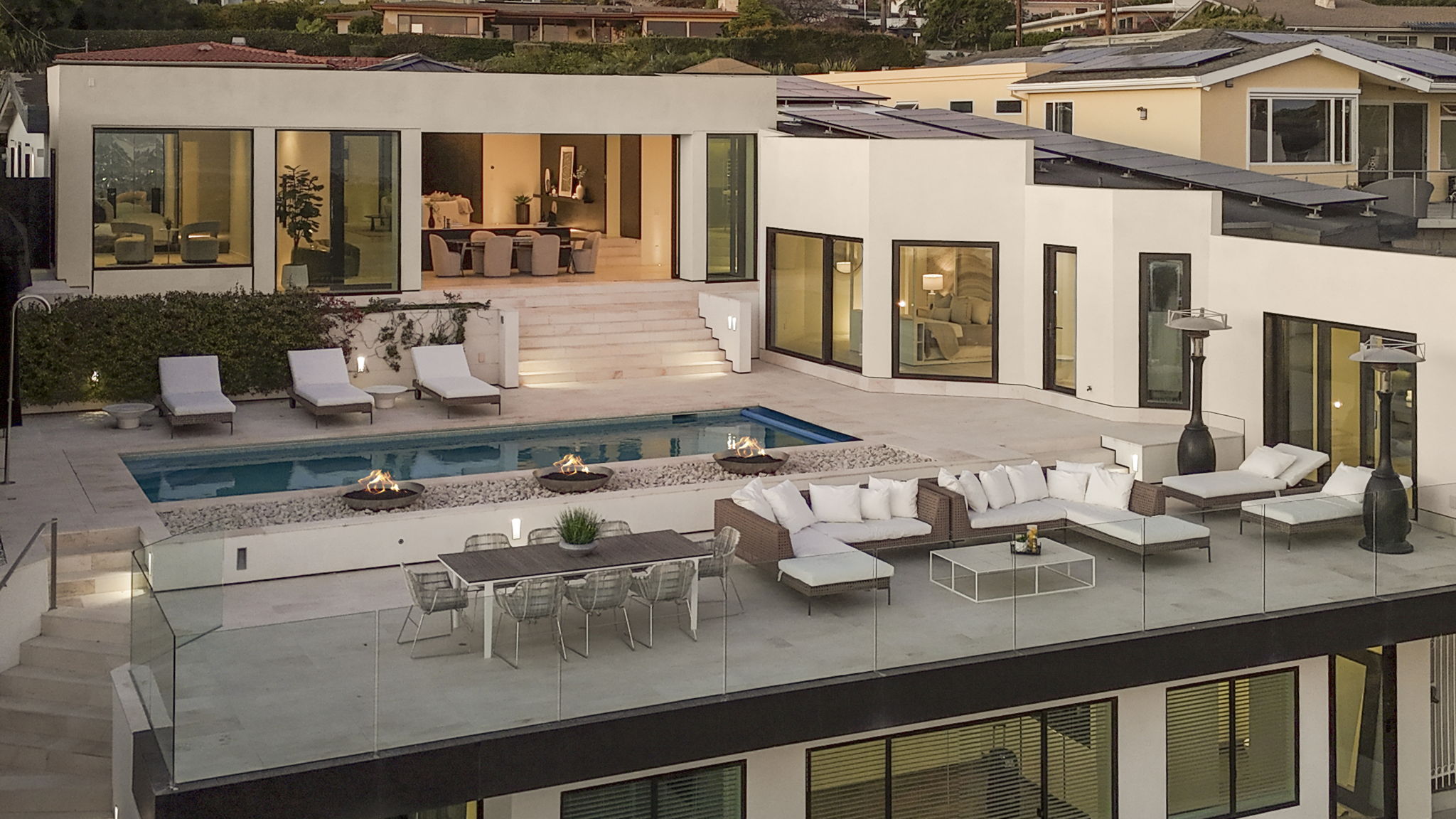 This modern luxury estate features a tiered outdoor living space complete with a pristine swimming pool and integrated fire pits. The sprawling patio includes multiple lounge areas with high-end modular furniture and a formal dining setup, all enclosed by a sleek glass railing. The architecture showcases clean lines, expansive windows, and a sophisticated aesthetic perfect for outdoor entertaining.