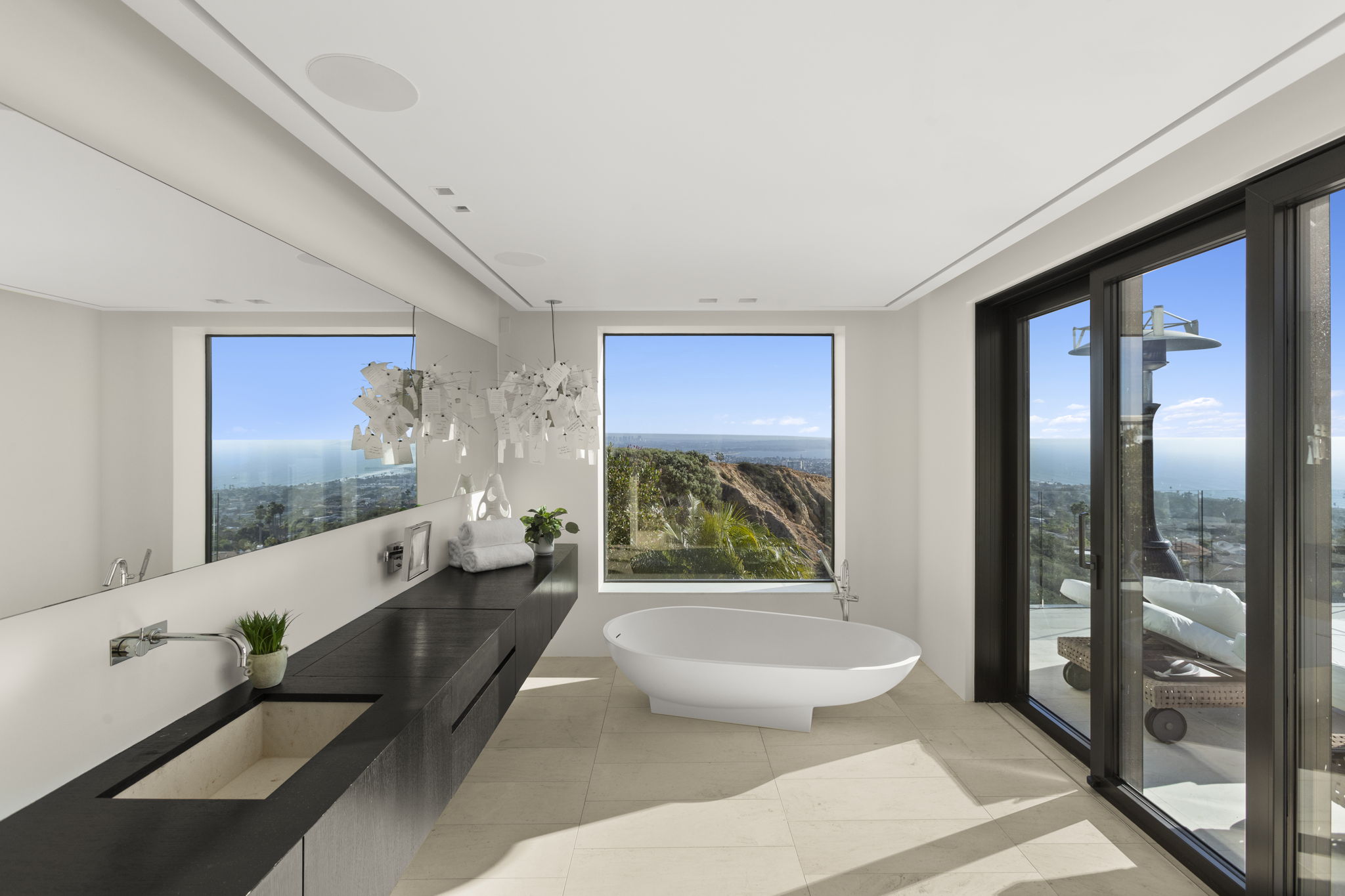 This modern, luxury primary bathroom features a sleek, minimalist design with a dark wooden vanity and integrated stone sink. A standalone bathtub is positioned centrally in front of a floor-to-ceiling window, which offers unobstructed, breathtaking ocean and scenic views. The space is further enhanced by an expansive mirror, high-end fixtures, and direct access to an outdoor deck, creating a bright and serene retreat.