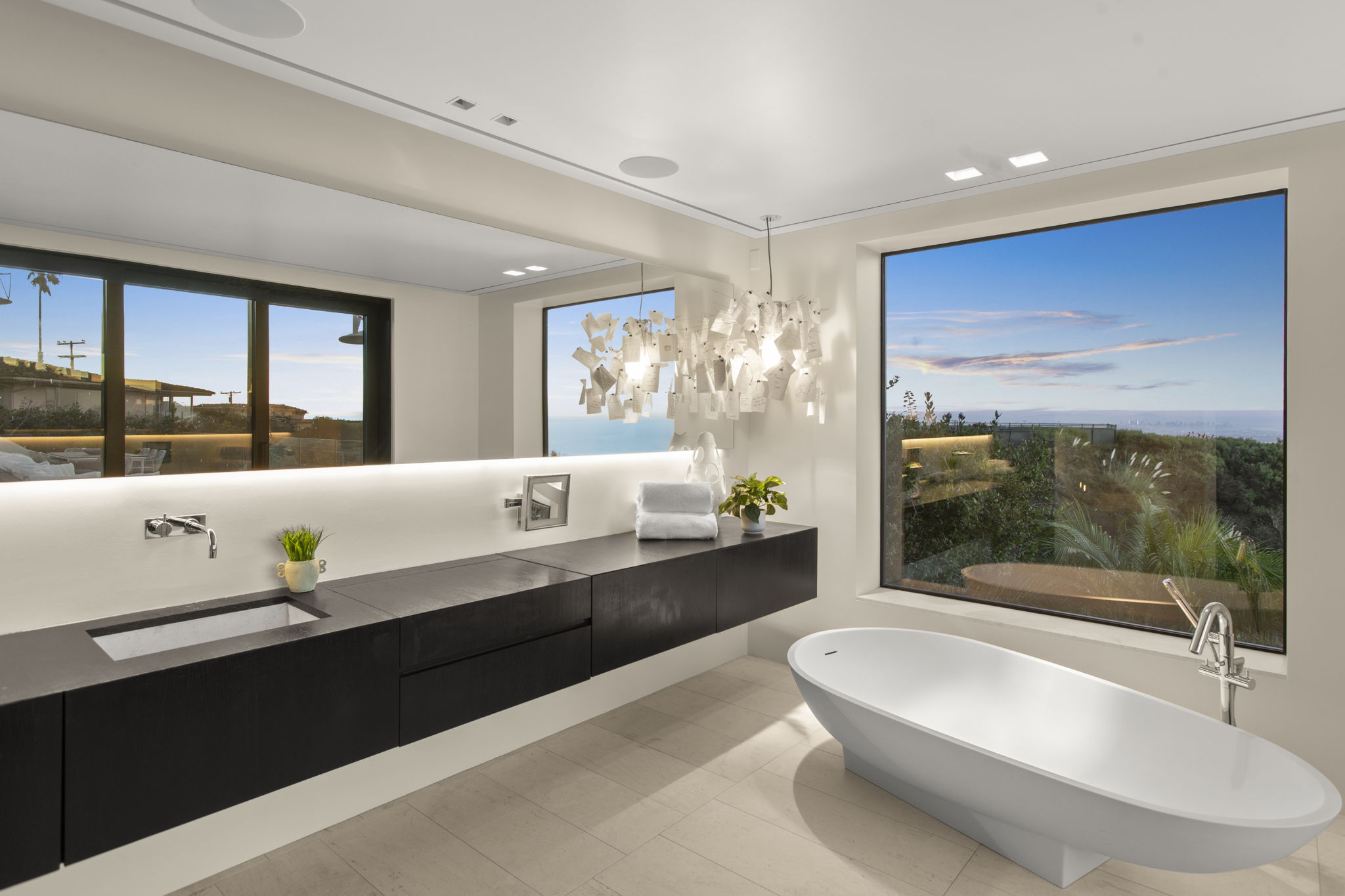 This modern, luxury primary bathroom features a floating dark wood vanity with a minimalist integrated sink and a large, full-width mirror with under-cabinet lighting. A sculptural chandelier hangs above, and a deep soaking tub is positioned by a expansive window offering scenic coastal views. The space exudes elegance and tranquility through its clean lines and sophisticated material palette.